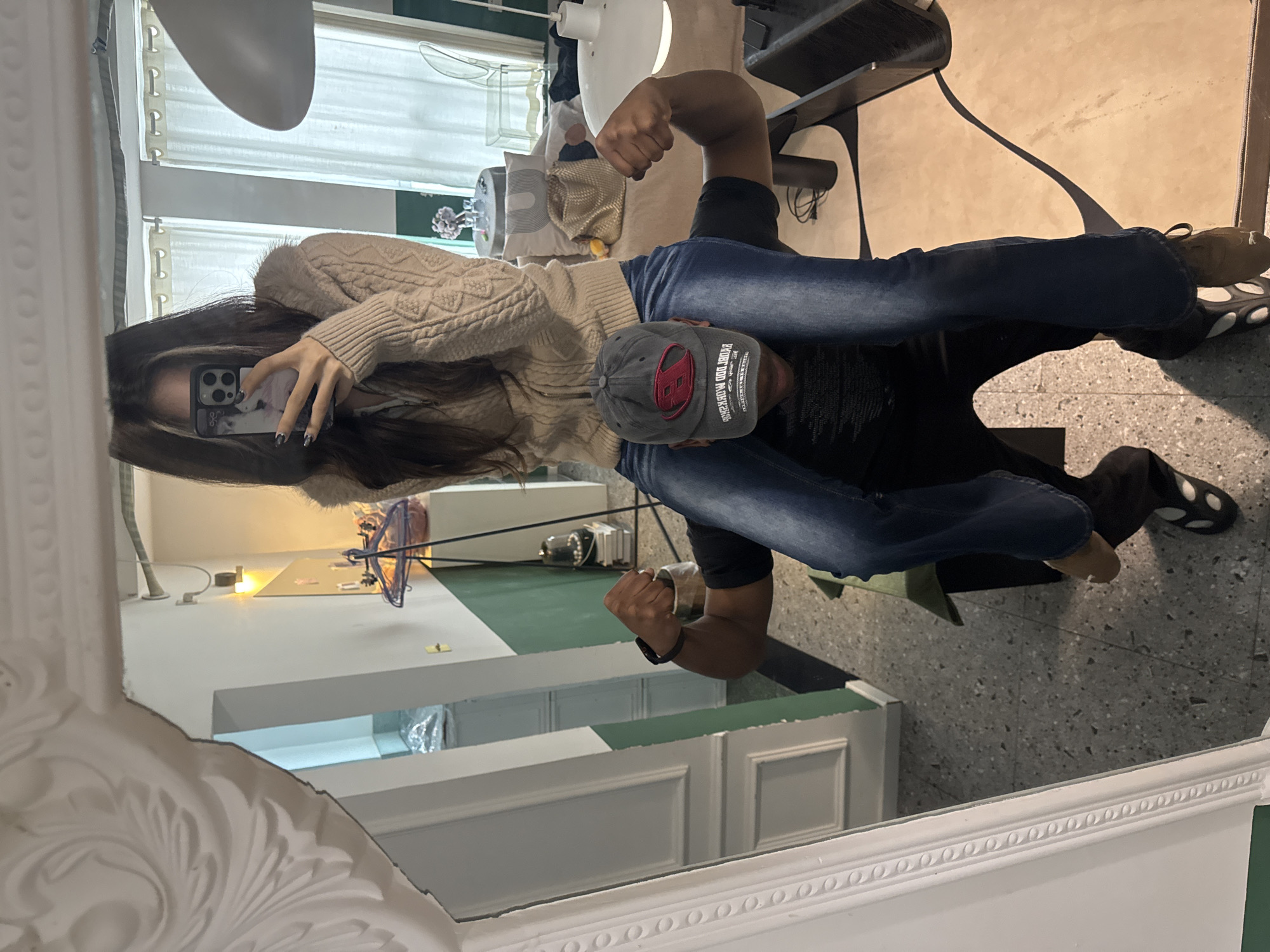 The image captures a mirror selfie of two individuals within an indoor setting, identified as located in Liangjiang, China. A female subject, with long dark hair and wearing a beige knitted cardigan over a lighter top and blue jeans, is positioned seated on the shoulders of a male subject. She holds a smartphone, featuring a dark case and visible camera array, towards the mirror to capture their reflection. Her fingernails are painted dark. The male subject, wearing a dark baseball cap with a red and black logo, a black t-shirt, dark trousers, and distinct black and white sandals, is on his knees, supporting the female, and is actively flexing his biceps with both arms. His face is obscured by the cap.
The scene is reflected in a large, white-framed mirror with ornate decorative moldings. The background elements visible in the reflection include a room with a light grey terrazzo floor and a two-tone wall, featuring a dark green lower section and a white upper section. To the left, a doorway with white trim is partially visible. To the right, a window with white curtains allows natural light into the space. A round, light-colored table is furnished with small glasses, a vase containing purple flowers, a decorative pillow, and a light-colored handbag. A transparent chair and a white pendant lamp are also present in the background.