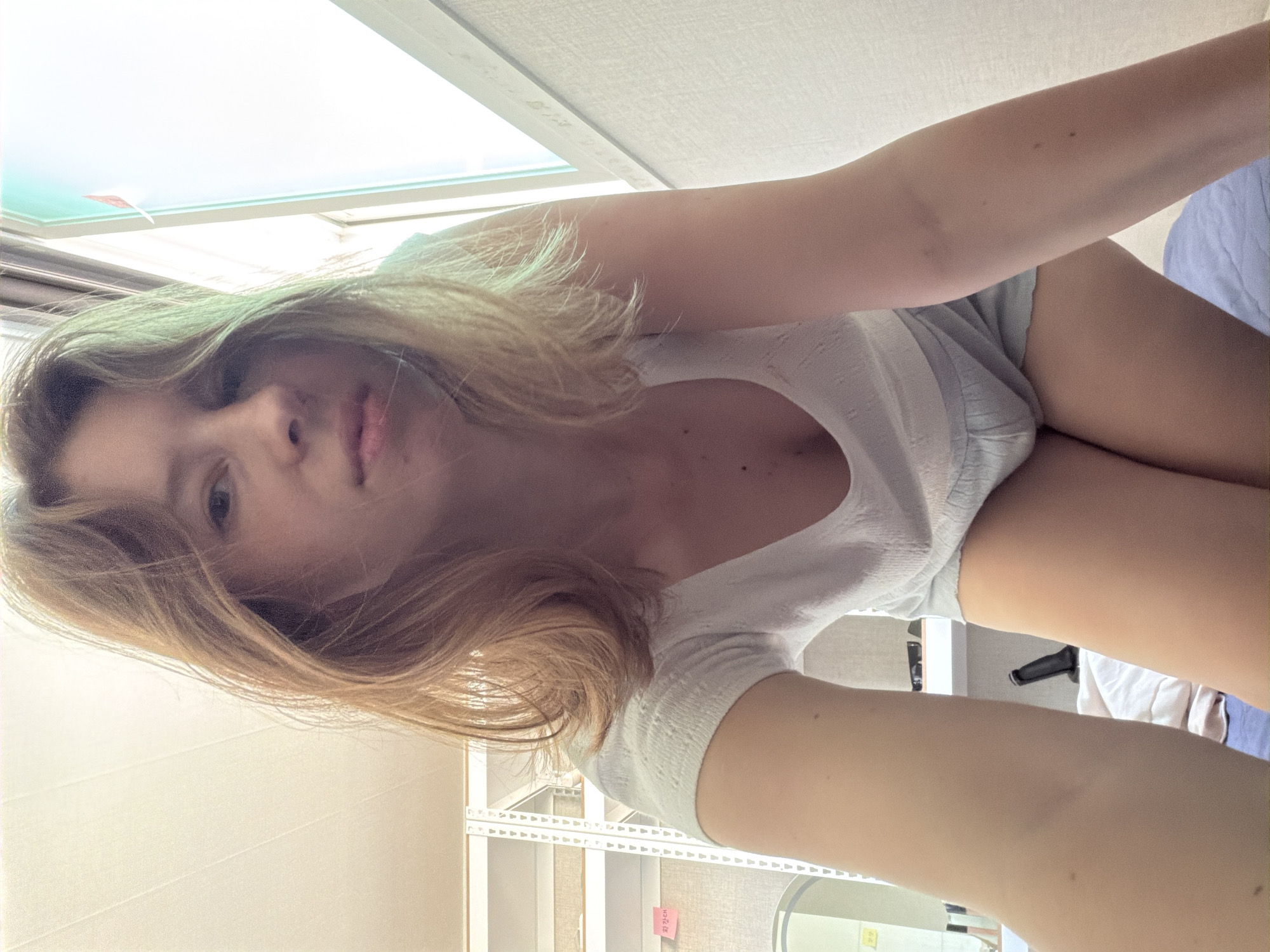 The media file features a close-up, sideways shot of a young woman with blonde hair. She is wearing a white t-shirt and light blue shorts. Her face is partially visible, and she appears to be looking towards the camera. Her arms and legs are also visible in the frame. The background suggests an indoor setting with shelving units and a window that lets in natural light, indicating it might be daytime. A small pink sticky note with Korean text is visible on one of the shelves. The overall mood is casual and candid. The location is identified as Dongducheon-si, South Korea.