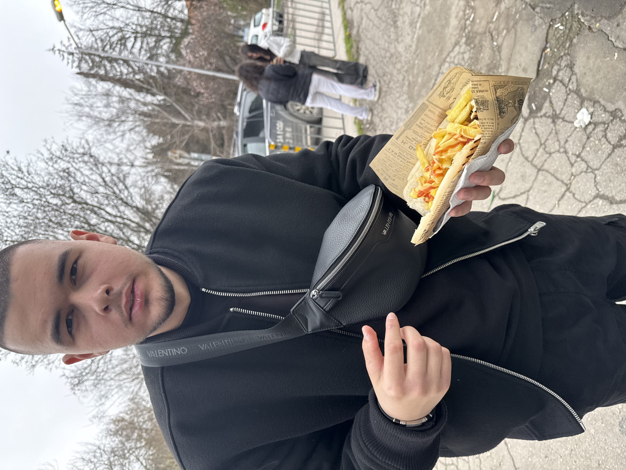 A young man stands outdoors, holding a paper cone filled with a sandwich overflowing with french fries and topped with a red sauce, likely ketchup. He wears a black jacket and a black Valentino crossbody bag. In the background, two people are seen standing near a car and a fence. Bare trees and a light pole are also visible. The ground appears to be cracked pavement. Based on the text in the food wrapper, it indicates it is a food establishment in Varna, Bulgaria.