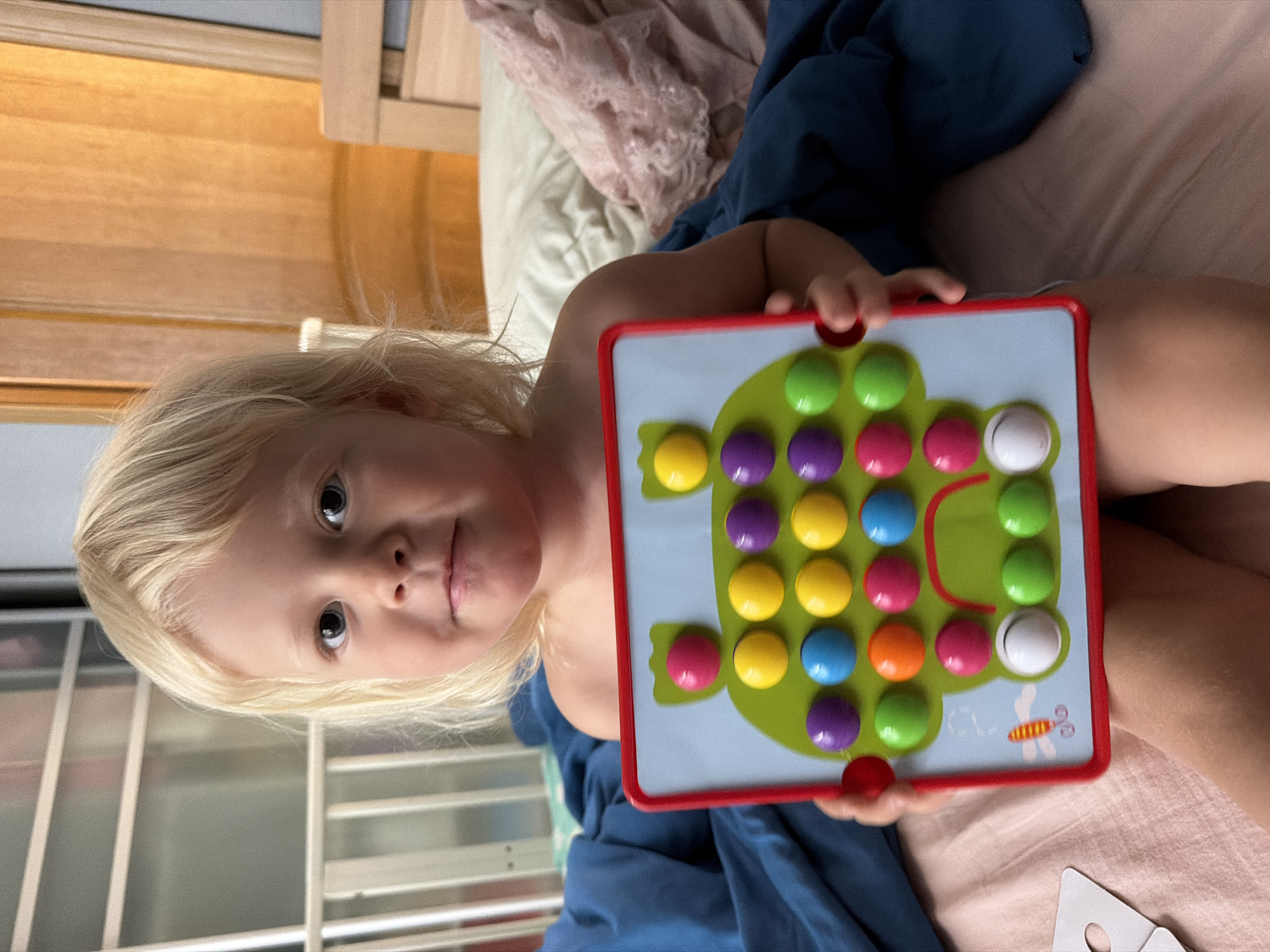 The image shows a young child with blonde hair holding a colorful pegboard toy. The child is looking towards the camera with a neutral expression. The pegboard features a green frog design and is filled with various colored pegs, including green, yellow, purple, blue, pink, orange, and white. The child is wearing a dark blue garment. The background suggests an indoor setting, possibly a bedroom, with wooden furniture and bedding visible. The lighting indicates it could be daytime. There is no visible text in the image. The location context provided is Sochi, Russia.