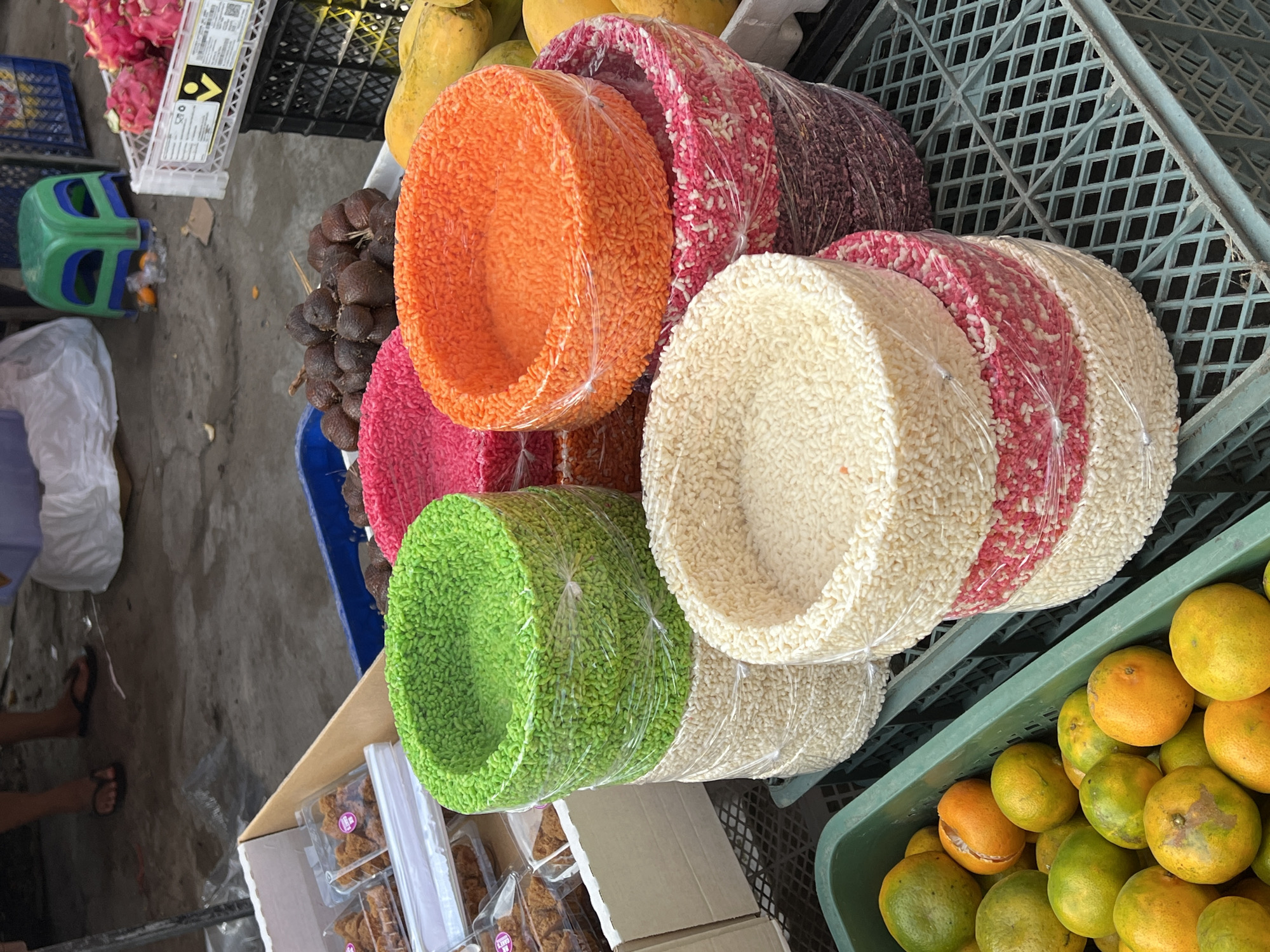 A close-up, eye-level, outdoor shot captures a vibrant market stall in Ubud District, Indonesia, overflowing with a colorful assortment of goods. In the foreground, a green plastic crate is filled with ripe oranges, their skins a mix of green and yellow. To the right, stacks of round, layered snacks wrapped in clear plastic are arranged on a blue plastic grid. The snacks feature concentric rings of various colors, including cream, pink, purple, green, and orange, each ring appearing to be made of small, puffed grains. In the background, several other items are visible: a pile of brown, scaly fruits, possibly salak, rests on a white surface. Further back, a green plastic stool is placed near a white fabric bundle, and to its right, a crate of bright pink dragon fruits and yellow papayas are displayed. The ground appears to be a rough, grey surface. Two individuals are partially visible in the upper left quadrant of the image: one stands with their legs visible, wearing dark flip-flops and a striped shirt, while another person is obscured behind a white bundle.