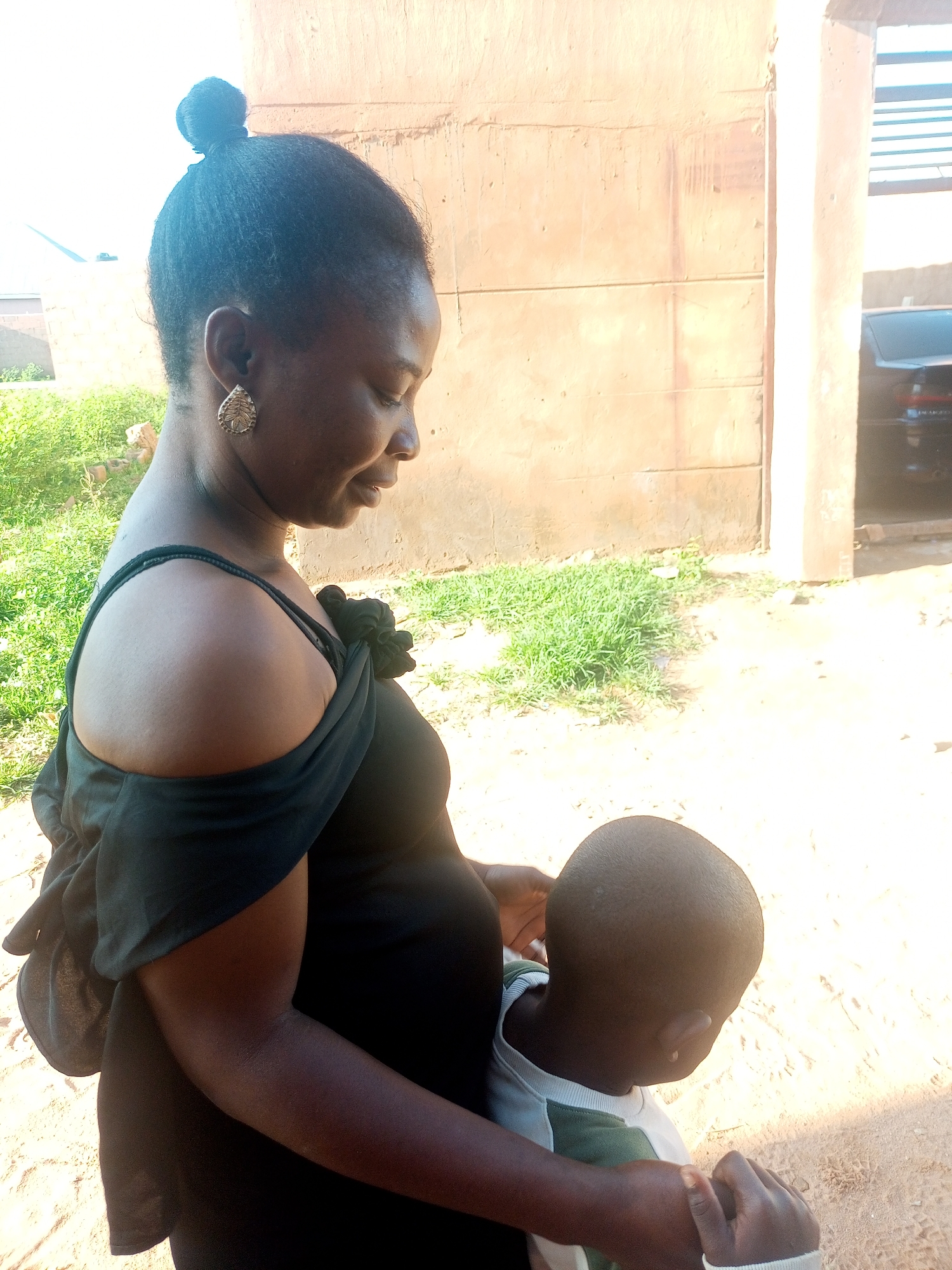 A pregnant woman in a black off-the-shoulder dress holds a young child who is facing away from the camera and has his head down. The woman has her hair in a bun and is wearing a large, decorative earring. The scene takes place outdoors in what appears to be a residential area in Tempure, Nigeria. The background includes a light-colored, textured wall and some sparse green vegetation. A portion of a dark vehicle is visible to the right of the frame. The ground is sandy.