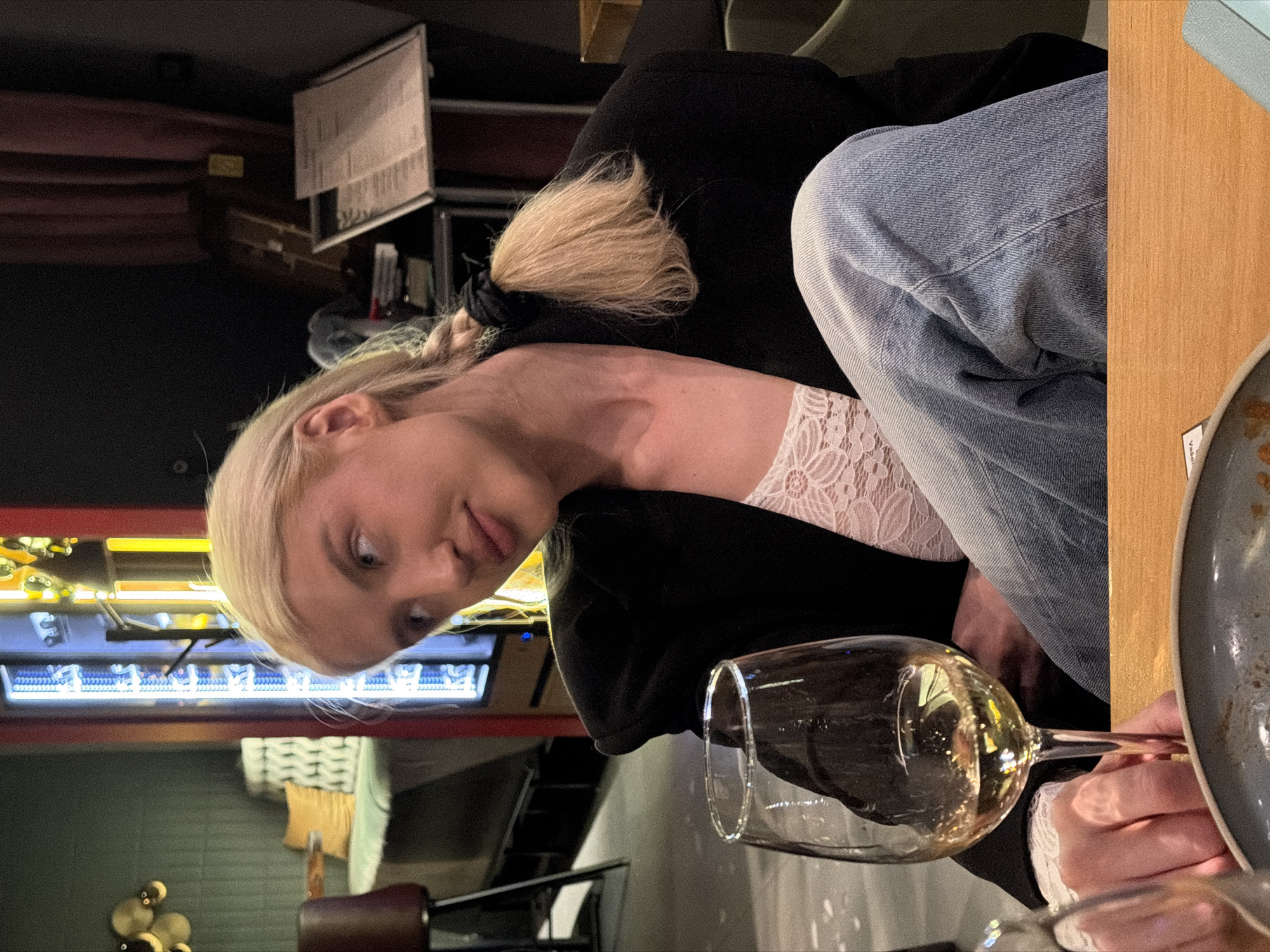 A blonde female is seated at a light-colored wooden table inside L'Atelier Bistrot & Bar, located at Kralja Petra 70, 11000 Belgrade, Serbia. Her hair is partially styled in a braid secured with a dark scrunchie, resting over her right shoulder. She wears a black blazer over a white lace top and light blue denim jeans. Her left hand holds a clear wine glass filled with a golden-yellow liquid, likely white wine. A dark grey ceramic plate is partially visible on the table in front of her. The subject's head is tilted slightly to the left, and she gazes upward and to the left of the frame. The background features a dim interior with various elements: to the left, a dark green wall with circular gold decor and a booth with patterned cushions and a yellow pillow; centrally, a vertical illuminated display column with blue/white light strips and a red frame, alongside other yellow neon lighting; and to the right, a dark wall section, a maroon curtain, and a silver-framed menu stand.