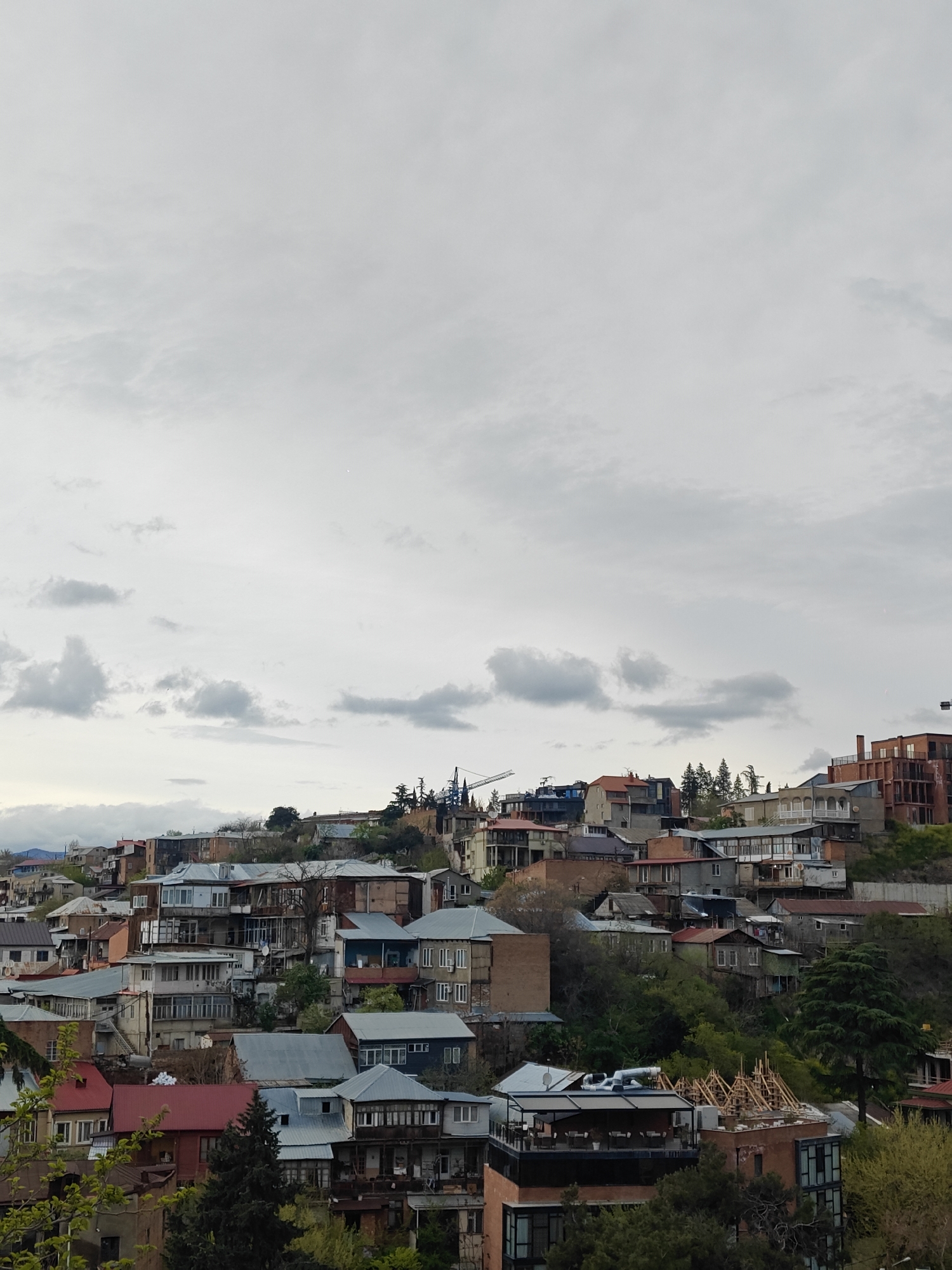 The image captures a panoramic view of a densely populated urban area built on a hillside, characteristic of Tbilisi, Georgia, under an overcast sky. The scene is dominated by a multitude of buildings, primarily residential, arranged closely together and ascending the slopes of the hill.
The architecture varies, showcasing a mix of older and newer structures with different roof types including corrugated metal, red tiles, and darker materials. Many buildings feature balconies, some enclosed, others open. Interspersed among the constructions are numerous green trees and bushes, clinging to the hillsides and dotting the landscape, providing pockets of natural greenery within the urban fabric.
The upper half of the frame is filled with a light grey, cloudy sky. Scattered cumulus clouds, slightly darker in tone, are visible, indicating an overcast daytime. The diffused light suggests a day without direct sunlight.
Evidence of ongoing construction and development is prominent. A tall yellow tower crane stands out against the sky near the center-left of the image, while another piece of construction equipment is visible further to the right. On a building in the lower right foreground, wooden scaffolding or roof framing is clearly visible, indicating active renovation or new construction. Some buildings appear to be in various stages of completion with exposed brickwork.
No people are visible in the image, nor is any legible text apparent on the buildings or signage. The photograph depicts a moment of an active, developing cityscape in Tbilisi.
