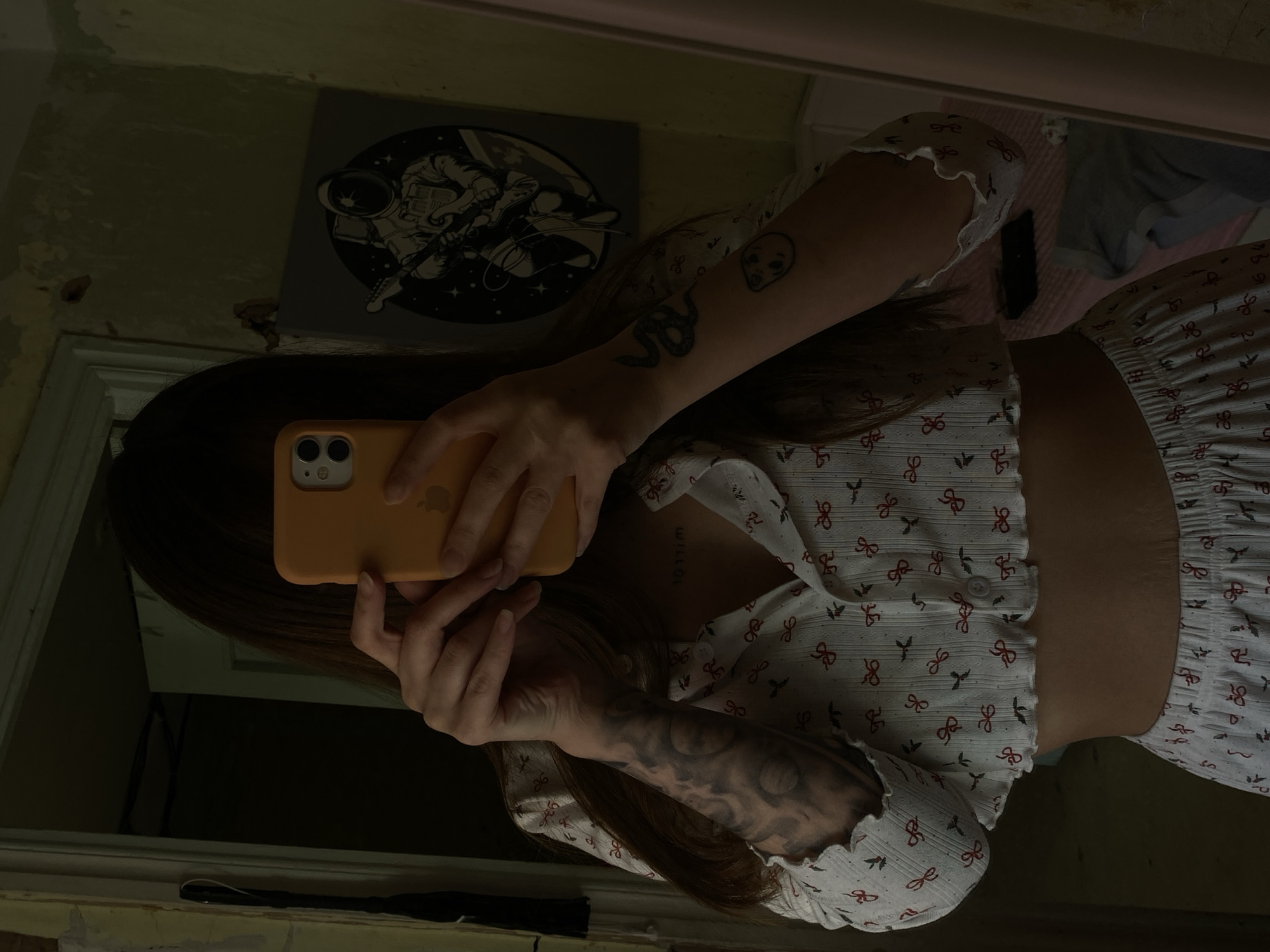 A person is reflected in a mirror while holding an orange smartphone to take a picture. The person has long hair and is wearing a white and red bow-patterned top and matching shorts. The person has multiple tattoos on their arms, including a snake and an alien face. Behind the person, there is a picture of an astronaut with a gun. The environment appears to be a room with faded paint and peeling walls, giving a slightly worn or aged impression. The location is not discernible from the provided image and is impossible to determine if it is in the city of Votkinsk, Russia.