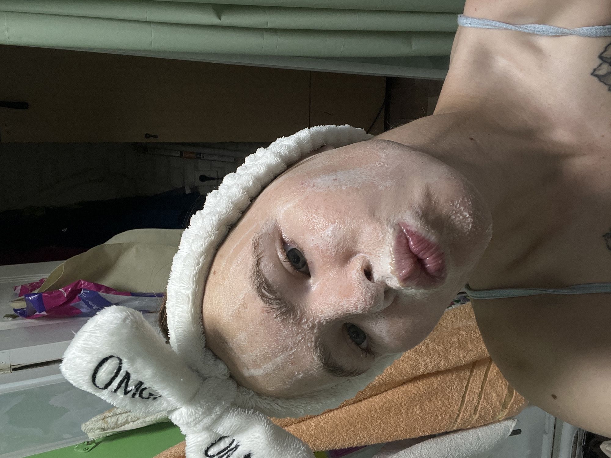 The image shows a young woman with a white fluffy headband with bunny ears on her head. Her face is covered in a white facial mask and she is making a duck face. She is wearing a light blue strap top. Behind her, there are towels hanging on a rack, a green shower curtain, and a door. The scene appears to be in a bathroom. There is no visible indication of the location in Chaikovsky, Russia.