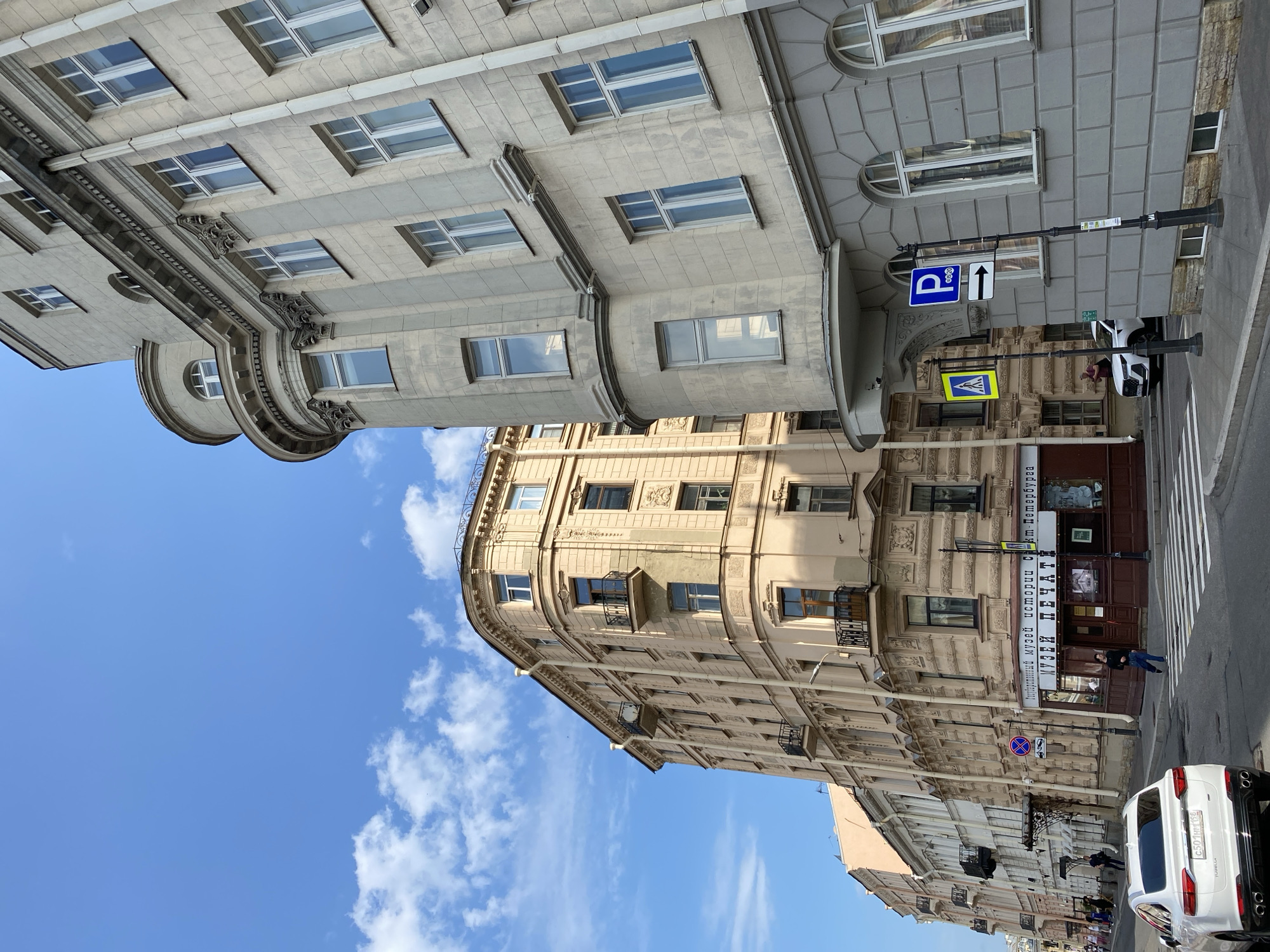 This is a daytime, eye-level, exterior, medium shot of a street corner in Saint Petersburg, Russia, focusing on two buildings with distinct architectural styles. The building on the right is a lighter gray, imposing structure with a rounded corner featuring multiple floors of windows and decorative molding. The building on the left is a pale orange-yellow, also multi-story, with balconies and ornate detailing, appearing older. A street intersection with a crosswalk is visible in the foreground, with a white SUV parked on the left side. Pedestrians are present, walking on the sidewalk and crossing the street. Signage includes a blue parking sign with an arrow and a yellow pedestrian crossing sign. The sky is bright blue with scattered white clouds. The environment appears to be a commercial or residential area with shops at street level, suggested by the "MUSEUM OF PRINT" signage on the orange building. The overall impression is one of a historic European city street.