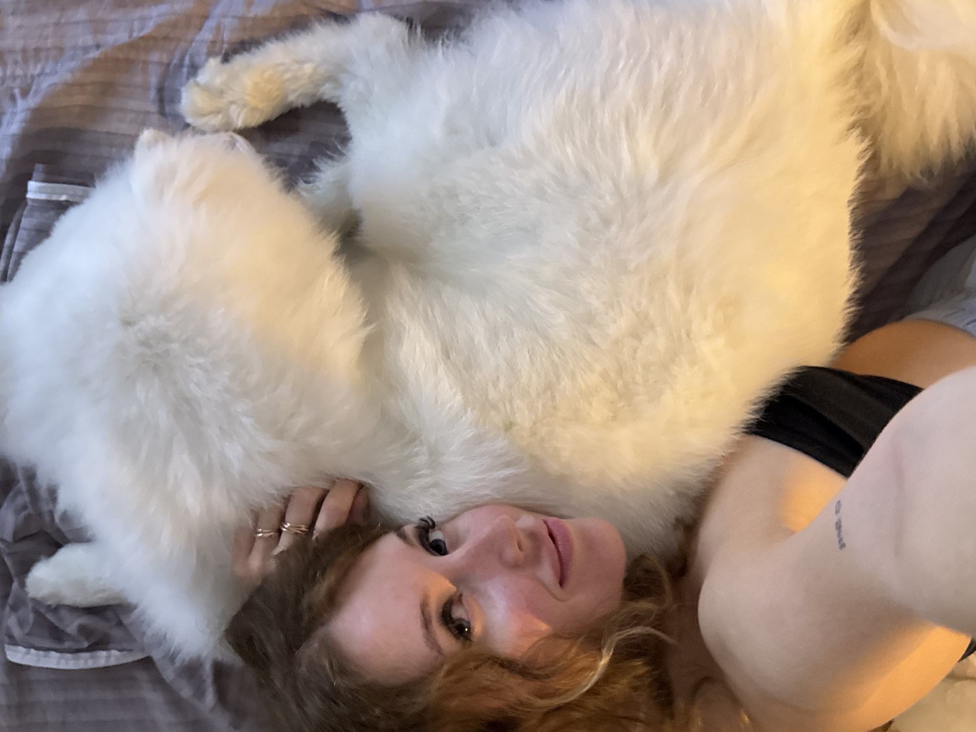 A high-angle medium shot shows a woman with curly auburn hair lying down next to a large, fluffy white dog. The woman is wearing a black top and has a tattoo on her left arm. She is looking up and smiling, with her right hand resting on the dog's head. The dog is mostly white with some yellowing fur on its paws. They are lying on a bed with a grey striped duvet. The image does not contain information about the city of Chaikovsky, Russia.