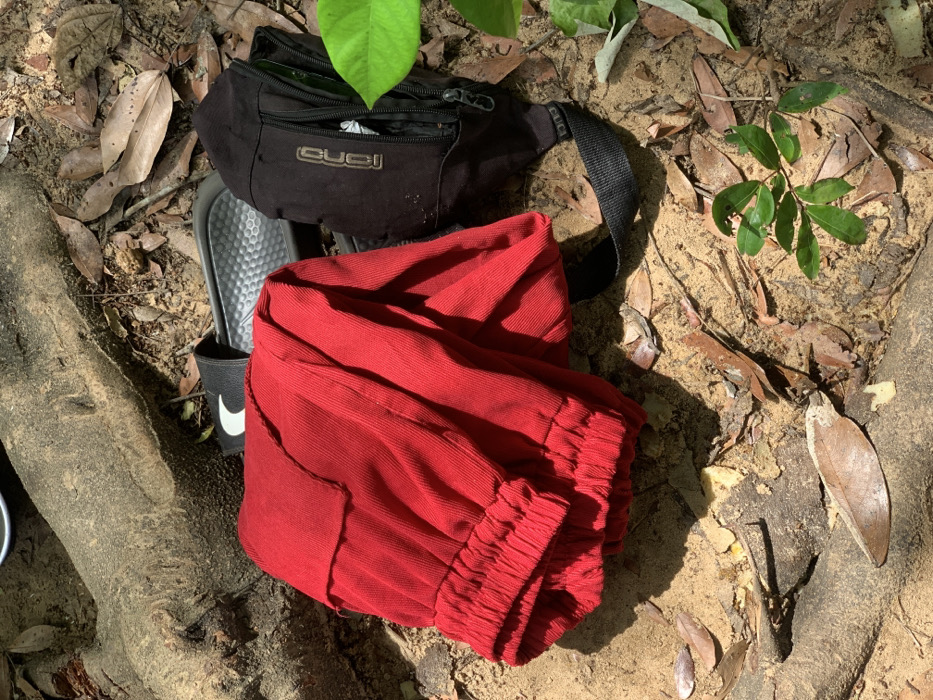 A pair of red corduroy pants are lying on the ground in Egbeda, Nigeria, near a black fanny pack and two pairs of flip-flops. The location is near the intersection of Olorunsogo Road and Ile-Ife Road, which is visible in the distance. The pants appear to be folded, and the fanny pack is laying on its side. The ground is covered in dry leaves and dirt, and the scene is lit by the sun. The fanny pack has the word "GUCCI" on the front, and the flip-flops are black with a white stripe. The environment is typical for a Nigerian city: dusty, with a mix of concrete and dirt. There are no people in the image. The scene is likely located in a residential or commercial area, based on the nearby buildings and street.