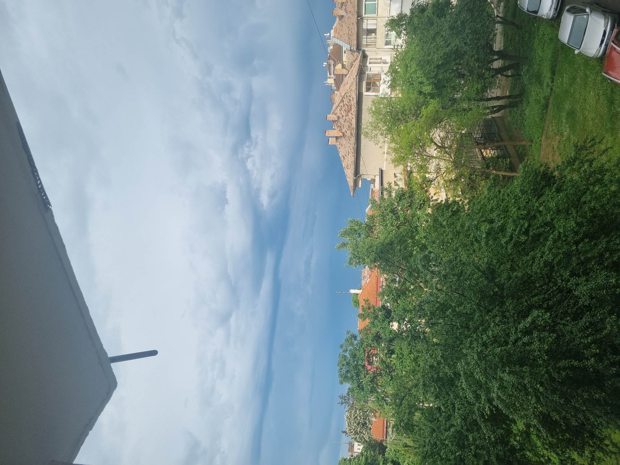View from a high point (likely a building), showing a street in Gorna Oryahovitsa, Bulgaria. A blue sky with wispy clouds occupies the left half of the image. The right half shows a building with orange tile roofs, interspersed with green trees. Parked cars are visible on a patch of green. The edge of the building from which the picture is taken is visible at the lower left.