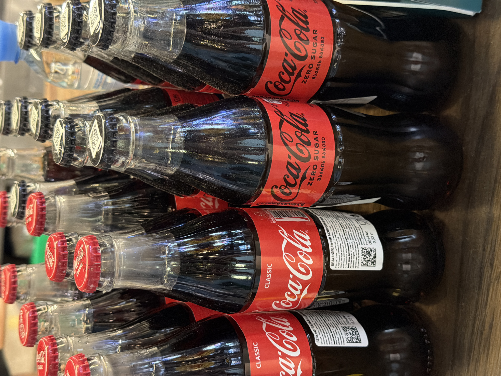 The image shows several glass bottles of Coca-Cola products stacked on a surface. The bottles are a mix of "Coca-Cola Classic" with a red cap and "Coca-Cola Zero Sugar" with a black cap. The text on the labels of the "Coca-Cola Zero Sugar" bottles appear to be written in Georgian, which suggests that the image may be taken in the country of Georgia, rather than Moscow, Russia. The bottles have QR codes on the label with ingredient information. The bottles appear to be in a store or storage area. The surface below the bottles looks like it's made of wood.