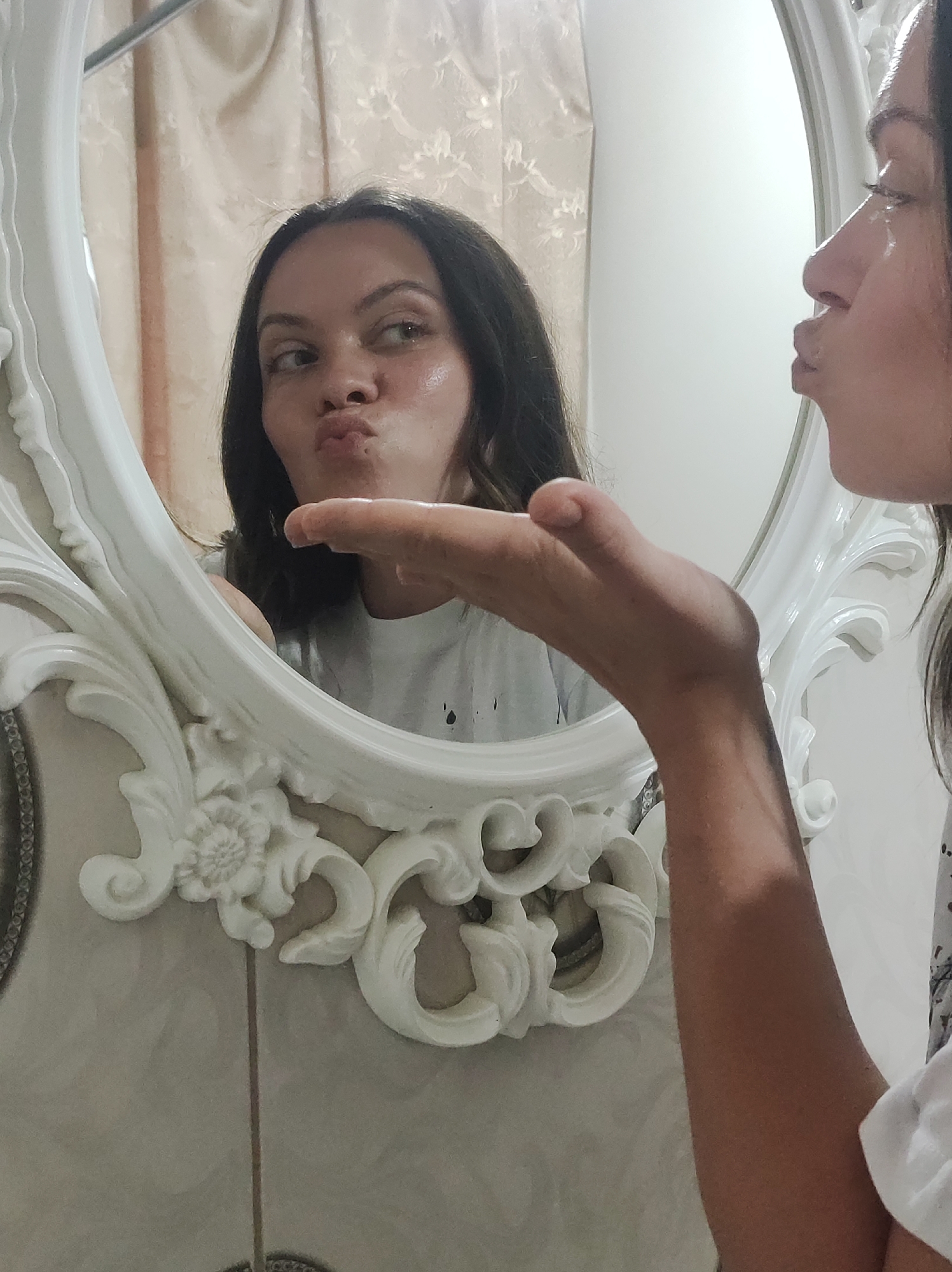 A woman is captured in a selfie taken in front of a large, ornate, white-framed mirror. The woman is facing the mirror, pouting her lips as if blowing a kiss. The mirror reflects her face, revealing dark hair and a white t-shirt. In the foreground, her arm is extended towards the camera, with her hand positioned as if releasing a kiss towards the viewer. The background behind the mirror consists of a tan-colored curtain and a light-colored tiled wall. The city location is Unknown, Unknown.