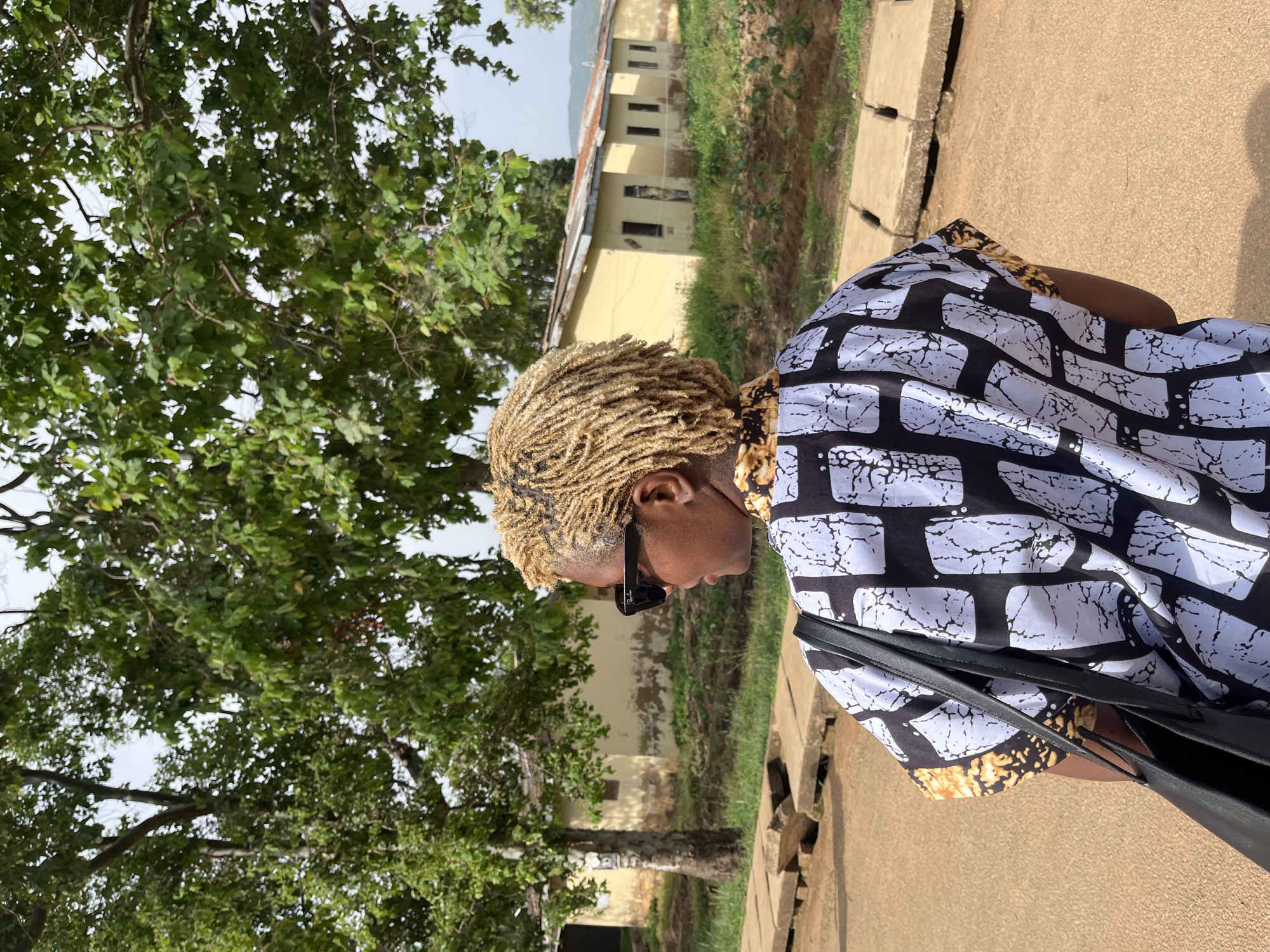 A person with blonde, dreadlocked hair and sunglasses stands facing away from the camera, looking to the left. They are wearing a black and white patterned shirt with yellow accents on the sleeves and carrying a black tote bag. The person is standing on a dirt path, with concrete slabs to their left. In the background, there are several small, yellow buildings with corrugated metal roofs, some of which appear dilapidated. Trees with green foliage are visible behind the buildings, and the sky is overcast. The scene is located in Rafin Par, Nigeria.