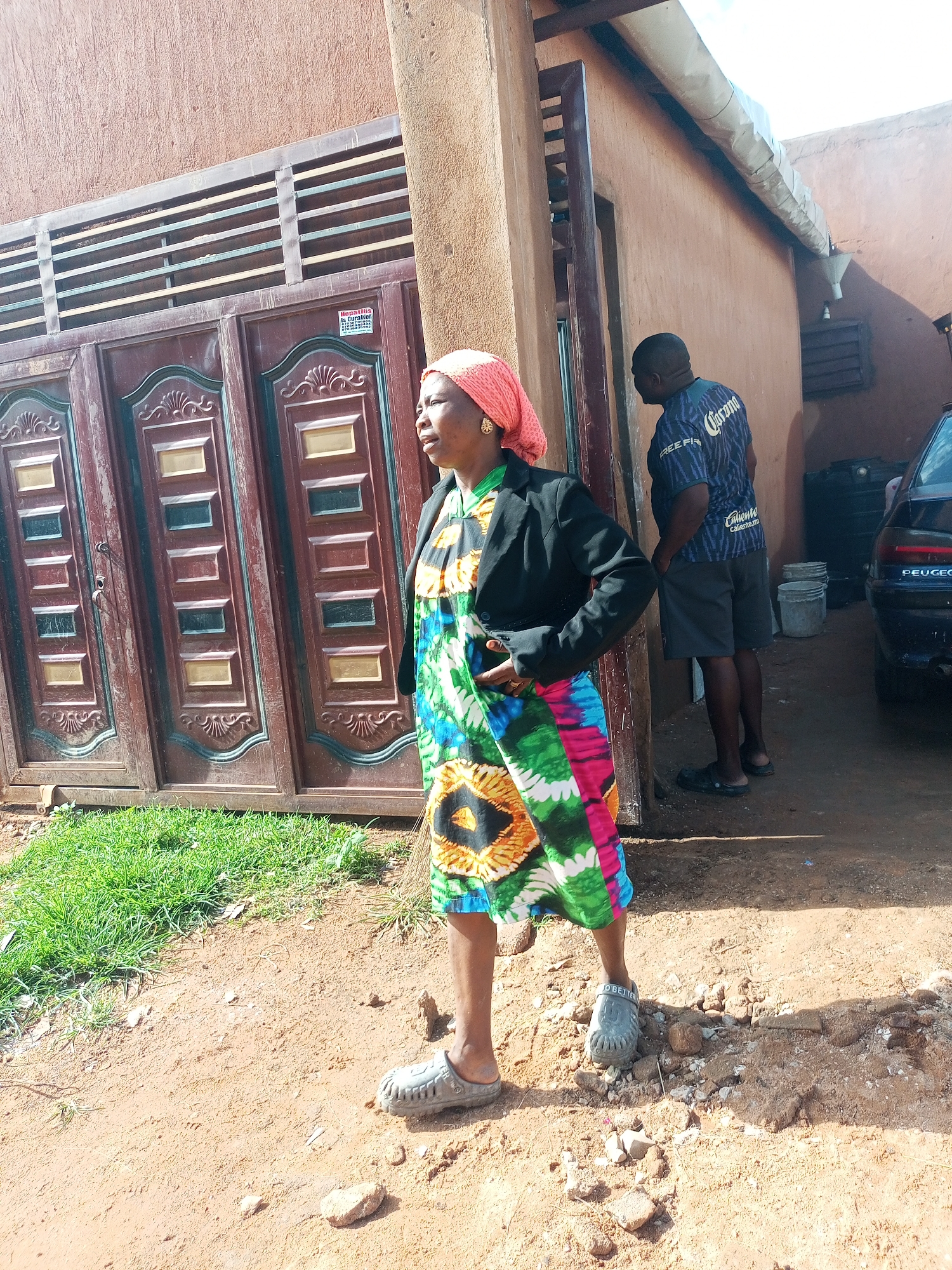 An outdoor, eye-level, medium shot captures a woman standing to the left of a large, ornate, red and gold metal gate. She is wearing a pink headscarf, a black jacket over a colorful, patterned dress, and gray clogs. Her right hand is tucked into her pocket, and she is looking to the left. Behind her, to the right, a man wearing a dark blue t-shirt and gray shorts stands with his hands in his pockets, facing away from the camera and towards a building. The building appears to be made of reddish-brown stucco and has a metal overhang. The ground is dirt with some patches of green grass on the left side. A black car is visible on the far right of the image, partially obscuring the building. The scene appears to be in a residential or commercial area.