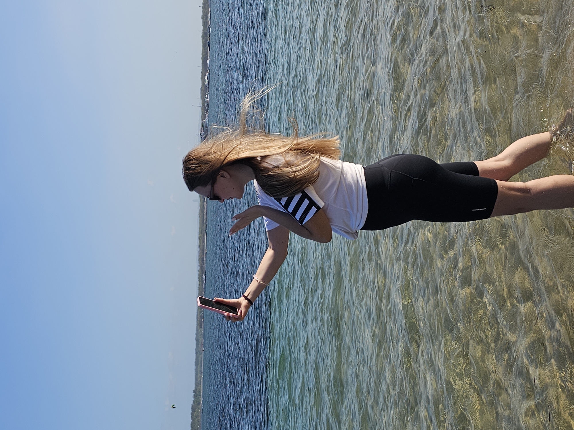 A young Caucasian woman with blonde hair is standing in shallow, clear water, taking a selfie with her pink smartphone. She is wearing sunglasses, a white t-shirt with black stripes on the sleeves, and black athletic shorts. Her right hand is holding the phone, while her left hand is near her face, appearing to blow a kiss. The background shows a body of water with a distant shoreline and a bright blue sky. Boats are visible on the water near the shore, and a kite surfer is in the distance. The scene suggests a tropical beach setting, possibly in the city of Denpasar, Indonesia, given the presence of ocean, boats, and a clear sky.