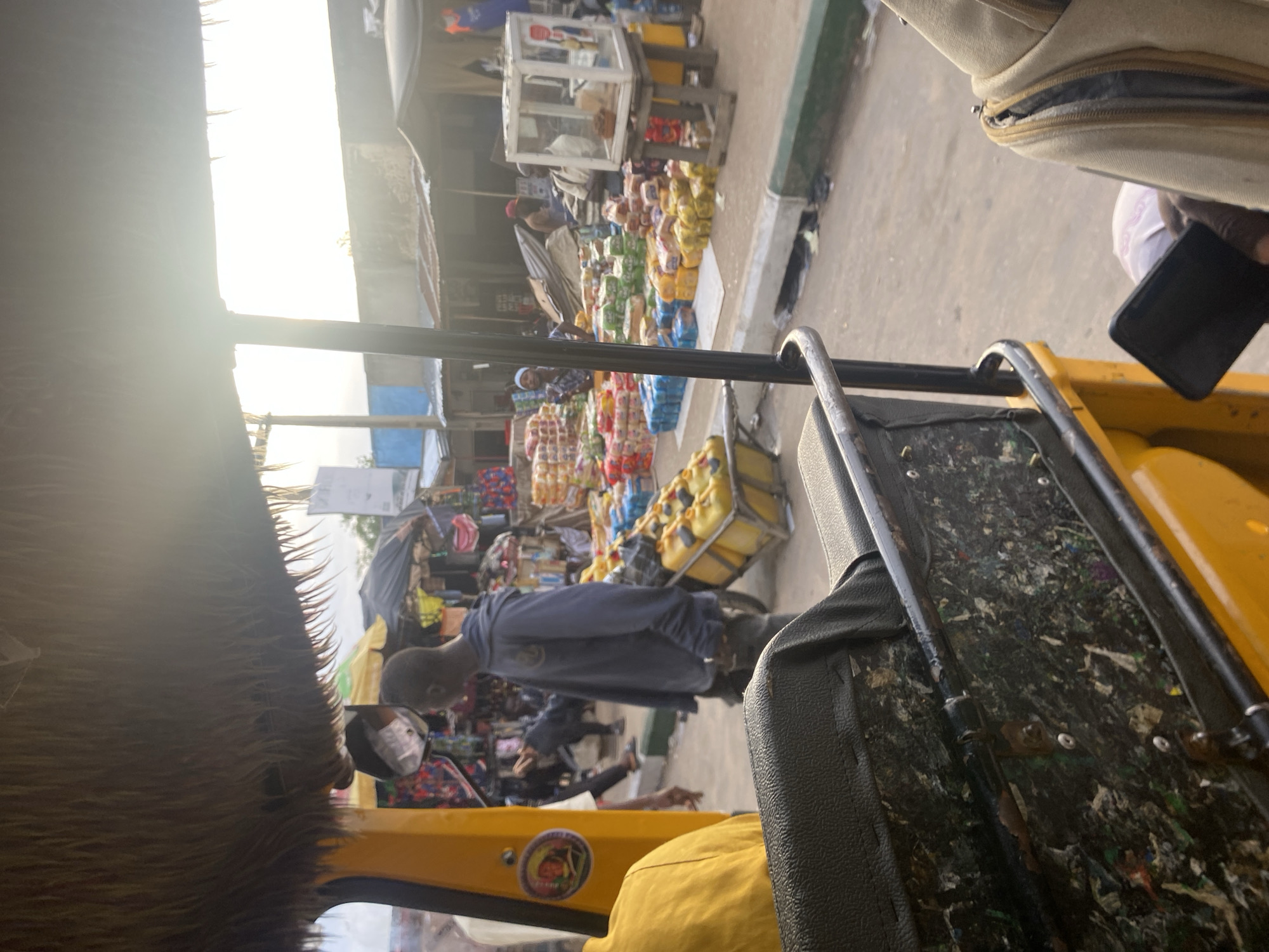 This image captures a bustling market scene in Jalingo, Nigeria, viewed from the interior of a yellow motorized rickshaw. The foreground shows the textured black seat and metallic railing of the rickshaw, with a colorful, somewhat distressed interior panel. Outside, a man in a blue hoodie stands near a cart loaded with yellow jerry cans, suggesting the sale or transport of liquids. The background reveals a vibrant marketplace with stalls overflowing with goods, including stacks of bread or packaged food items in various colors. Several other people are visible, engaged in market activities, with awnings and makeshift roofs providing shade. The overall atmosphere is one of lively commerce under the bright, possibly late afternoon sun.