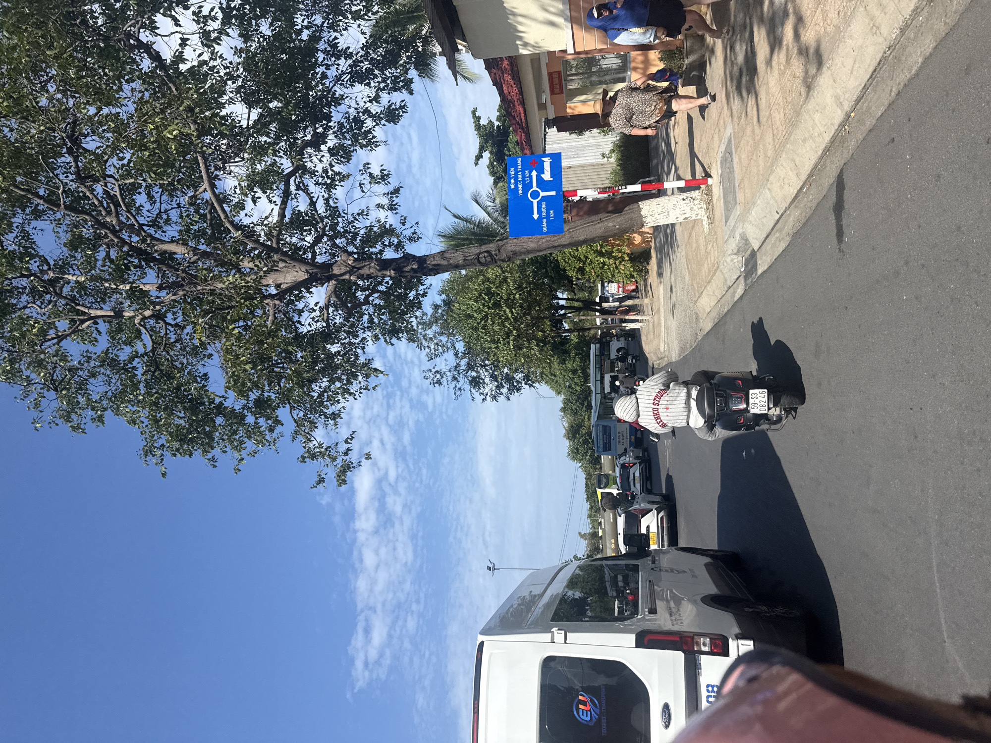 The image depicts a street scene in Nha Trang, Vietnam, under a bright, clear blue sky with scattered clouds. The perspective is from a vehicle, looking forward into traffic. A white passenger van with a blue and red "ELi" logo and "TOURIST TRANSPORT" text is on the left. In the center lane, a person on a black scooter, wearing a white hooded jacket with red and white stripes and "MENKO STUDIO" text, is ahead, moving away from the camera. The scooter has a license plate that reads "59-S3 08246". Several cars and buses are visible in the background, suggesting moderate traffic.
To the right of the road, a blue directional sign with white text and symbols is attached to a tree with a red and white striped pole. The sign indicates directions to "BỆNH VIỆN VINMEC NHA TRANG 1.2 KM" (Vinmec Nha Trang Hospital) and "QUẢNG TRƯỜNG 1 KM" (Square). Adjacent to the sidewalk, there's a building with an orange and yellow facade and a tiled roof. Two women are walking on the sidewalk; one is wearing a patterned dress and a straw hat, and the other is wearing a blue headscarf and a blue top with dark shorts. The sidewalk is lined with trees and planters. The overall environment appears to be a sunny day on a well-maintained urban road.