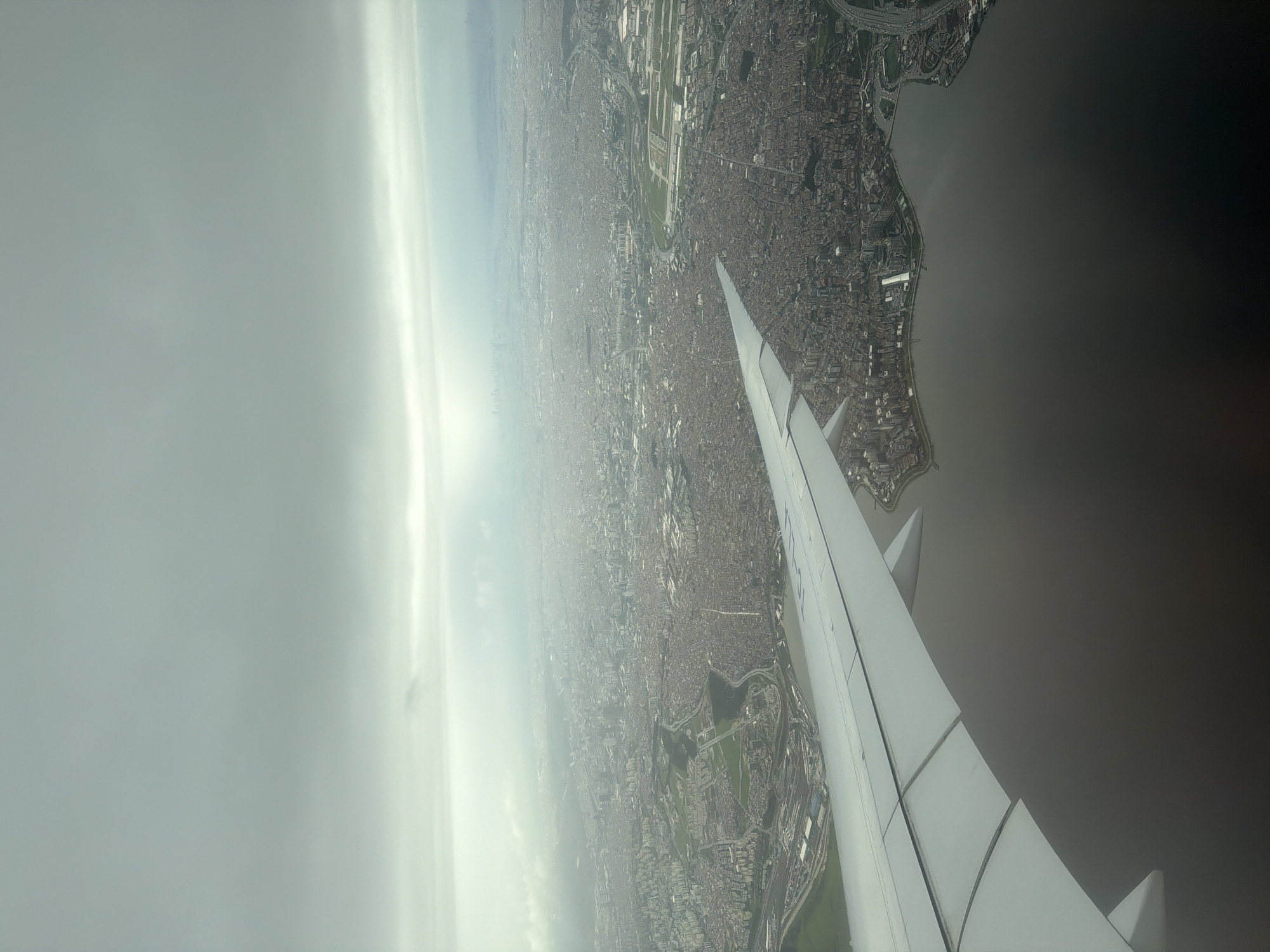 An aerial view from an airplane window shows a section of Arnavutköy, Turkey, under hazy skies. The foreground contains the wing of the plane. Below, dense urban development stretches from the coastline inland. The city appears to be built on a grid pattern, with buildings densely packed together. Roads and highways weave through the urban fabric, and some open green spaces can be seen. The body of water adjacent to the city is dark. The sun is obscured by clouds, creating a soft lighting effect.