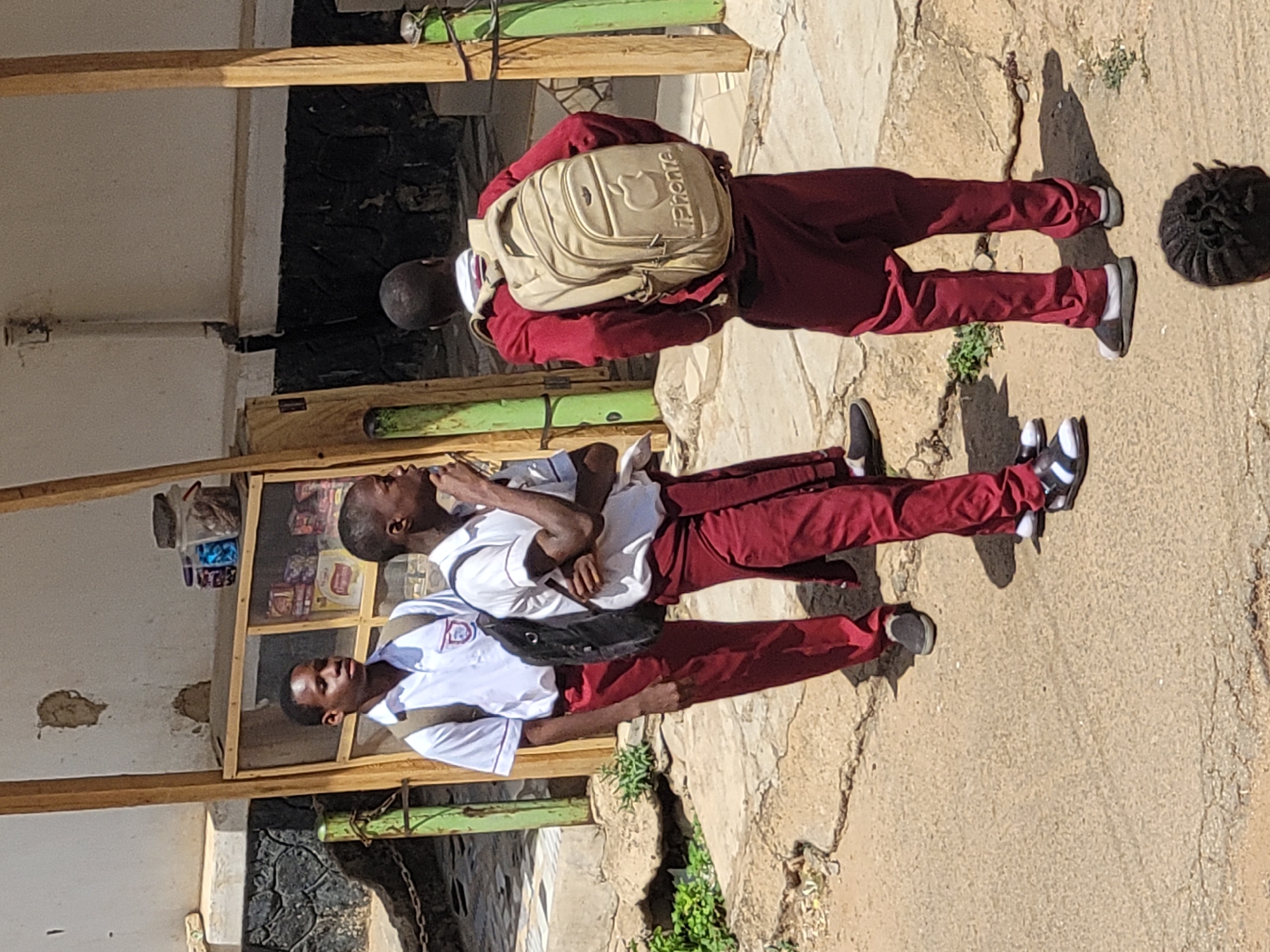 Three African male students in maroon trousers and white collared shirts are standing on a paved surface in front of a building in Jos, Nigeria. The student closest to the camera wears a black shoulder bag. The student in the middle has crossed arms. The student furthest away wears a beige backpack. A small store with items for sale is visible behind the students. The environment includes green vegetation and the building has wooden support structures.