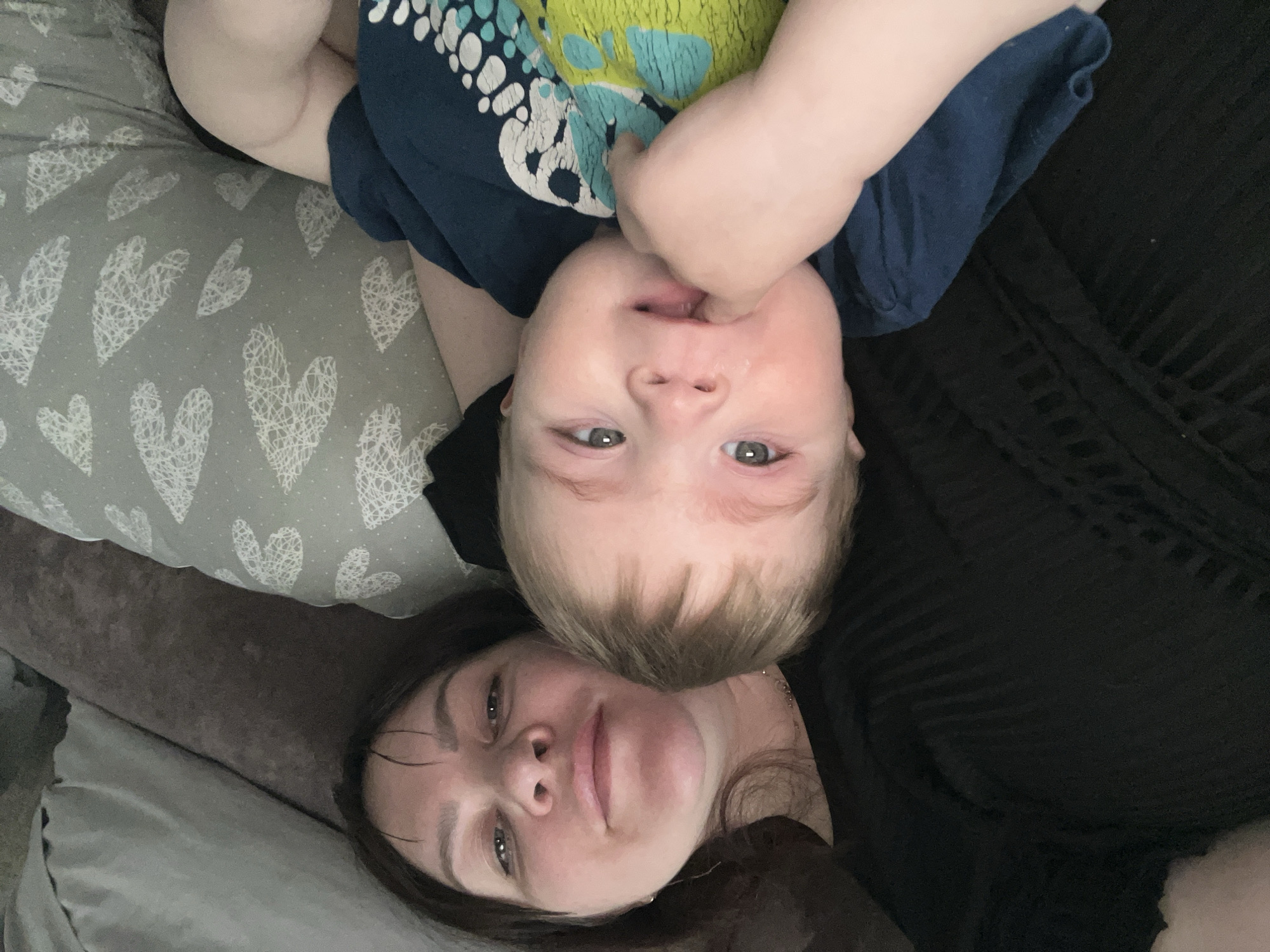 A selfie-style photograph shows a woman and a young child lying down. The woman is at the bottom of the frame, lying on her back, with her face close to the camera. The child is positioned above her, also lying on their back, but with their head oriented towards the top of the frame. The child is wearing a dark blue t-shirt with a colorful graphic on the front. Their hand is raised towards their mouth. The woman is wearing a dark-colored shirt. The background includes a pillow with a white heart pattern and a dark-colored couch. The location and city of Noviy, Russia are not possible to determine from the image.