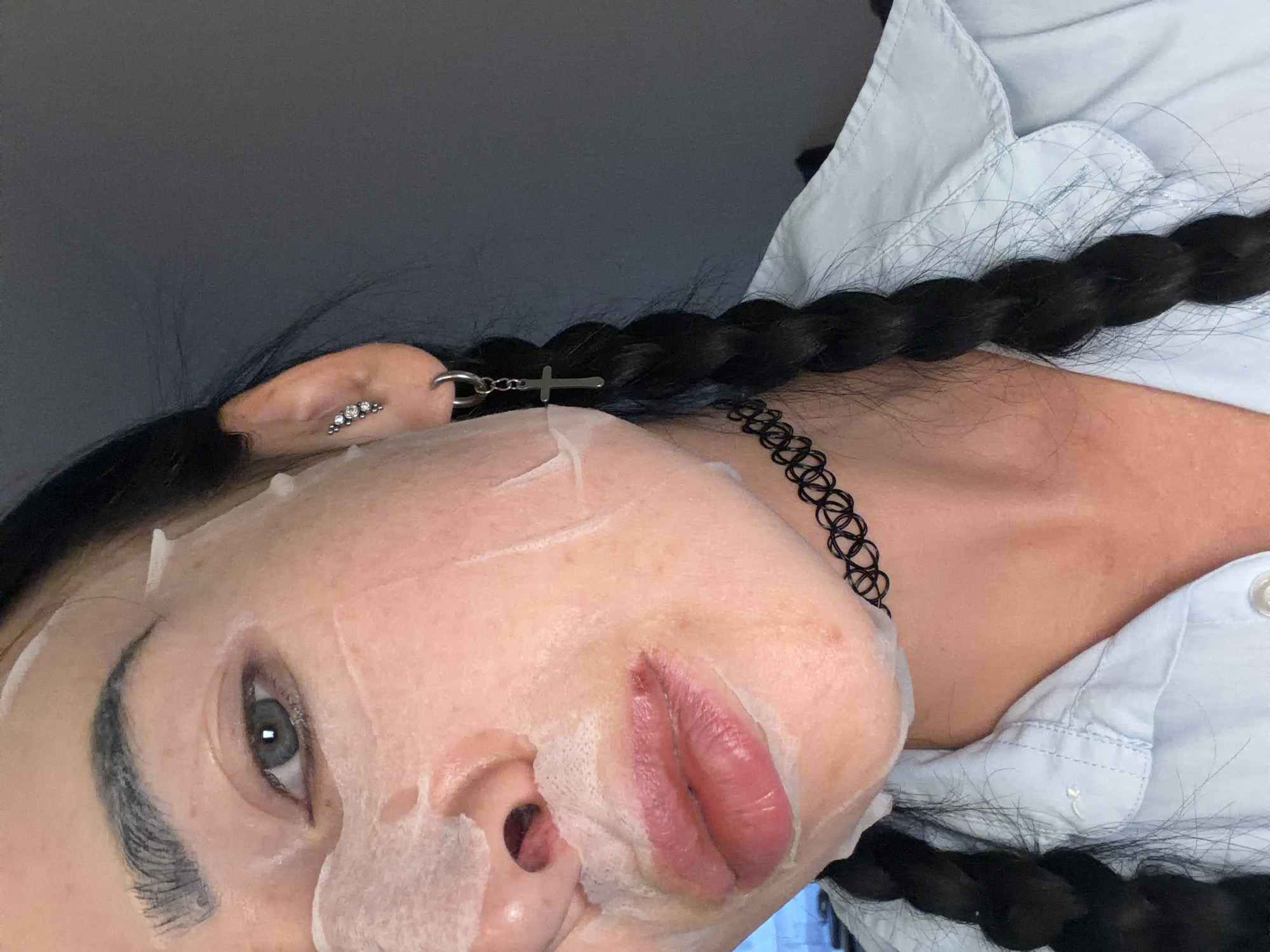 The image shows a close-up of a person's face, with a sheet mask applied. The person has dark hair in two braids and is wearing a light blue collared shirt, a black choker necklace, and earrings. One earring is a stud with multiple small stones, and the other is a hoop with a cross charm. The sheet mask covers most of the face, obscuring the nose and forehead, and extends down to the chin. The person's lips are visible and appear to be painted with a pink lipstick. The background is dark and out of focus, making it impossible to determine the location or setting. There are no discernible elements that would indicate the image was taken in a specific city or country.