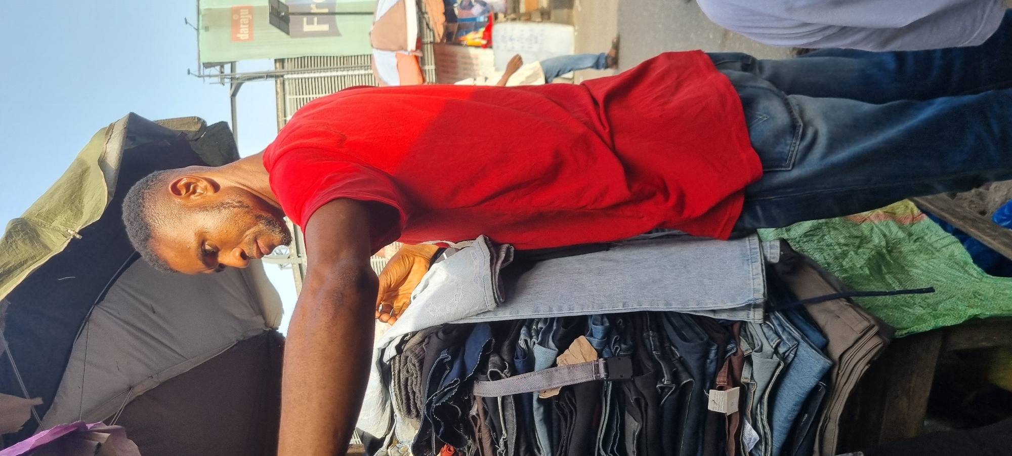 A street vendor in Lagos, Nigeria, stands behind a stack of folded and hanging clothing. He is a dark-skinned man wearing a red shirt and jeans. He is bent forward, looking at the merchandise. The stack of clothing is a mix of jeans and other garments. Behind him is a makeshift tent with a green and brown tarp. In the background, there are buildings and people, including someone wearing a white shirt and jeans. A billboard is visible with the word "Daraju."