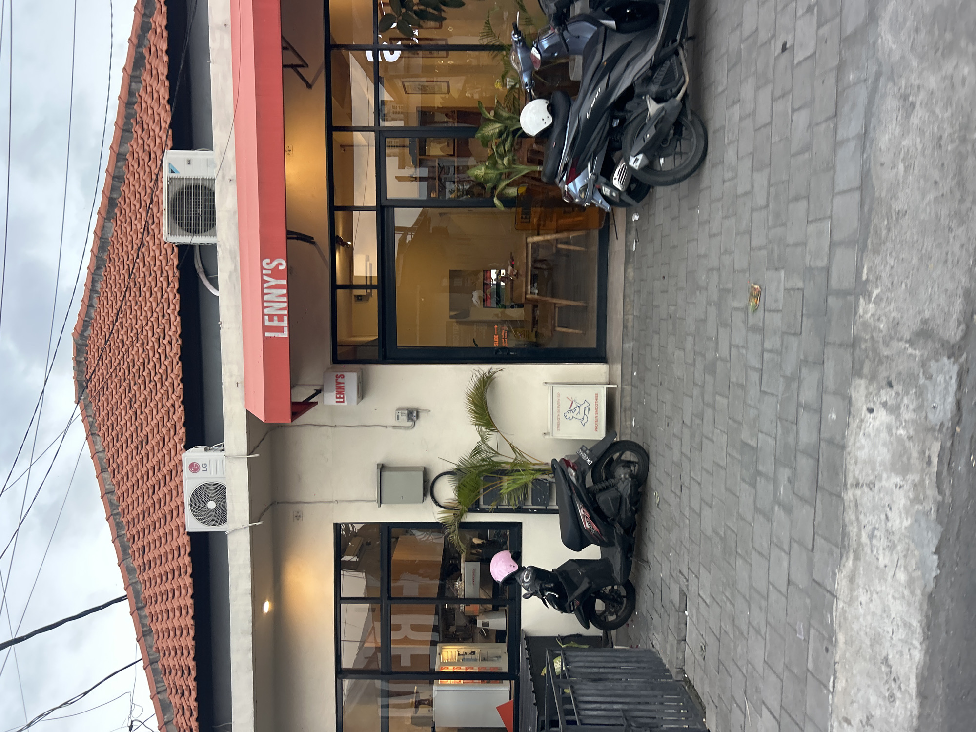 The image shows the exterior of a cafe in North Kuta, Indonesia. The cafe has a red awning with the name "LENNY'S" printed in white letters. In front of the cafe, there are two scooters parked on a paved sidewalk. The cafe has large glass windows, allowing a view into the interior where tables and chairs are visible. Several potted plants are placed outside the cafe. An air conditioning unit is mounted on the exterior wall above the awning. Overhead, power lines are visible against an overcast sky.