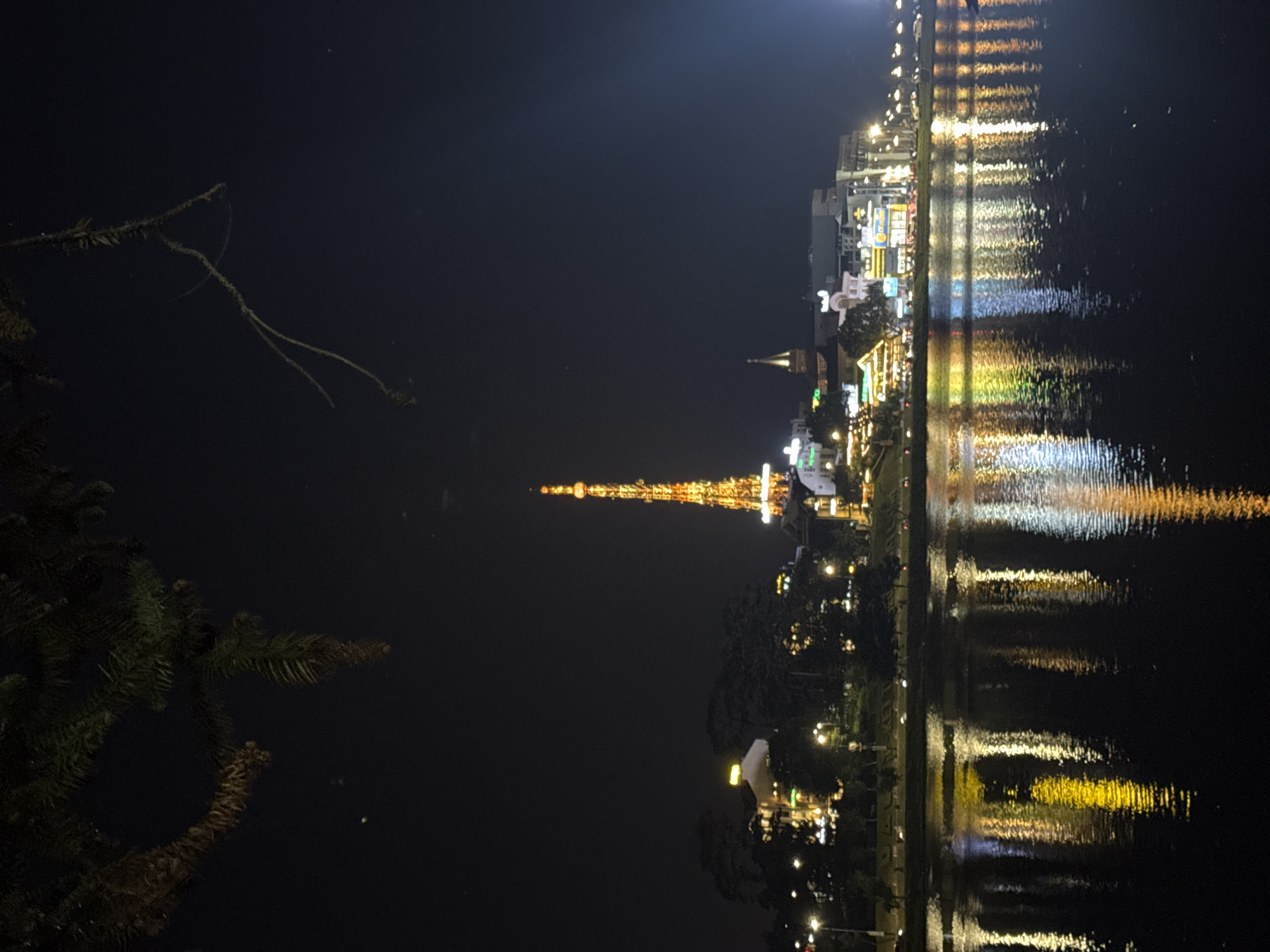 The image depicts a nighttime scene in Da Lat, Vietnam, featuring Xuan Huong Lake. The iconic Da Lat Eiffel Tower replica is illuminated, along with nearby buildings and structures. Reflections of these lights are visible on the surface of the lake. There is vegetation, possibly trees, silhouetted on the left side of the image.