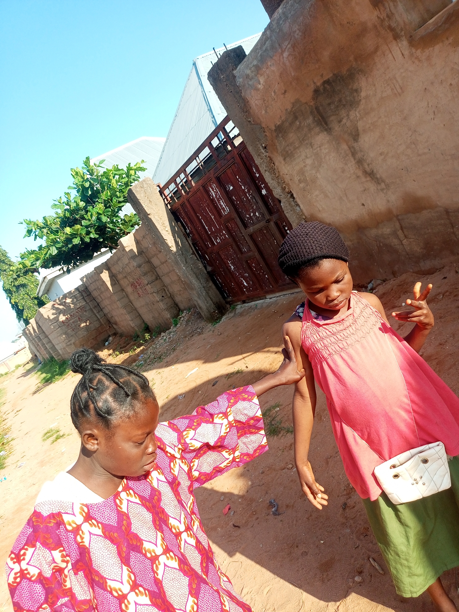 In Tempure, Nigeria, two young girls are captured outdoors against a backdrop of a red metal gate, a concrete wall, and a clear blue sky. The girl on the left is wearing a vibrant pink and white patterned dress with a white undershirt. Her hair is styled in braids pulled into a bun. She is looking down at something in front of her, with her left arm extended towards the girl on the right, her hand resting on the other girl's shoulder. The girl on the right wears a dark brown knit cap, a coral pink sleeveless dress, and a green skirt. A small white quilted bag with gold accents is attached to her hip. She is looking downwards, with her right hand raised, fingers spread, as if in a gesture. The ground is a mix of sand and dirt, with some sparse vegetation. The environment suggests a casual, everyday setting.