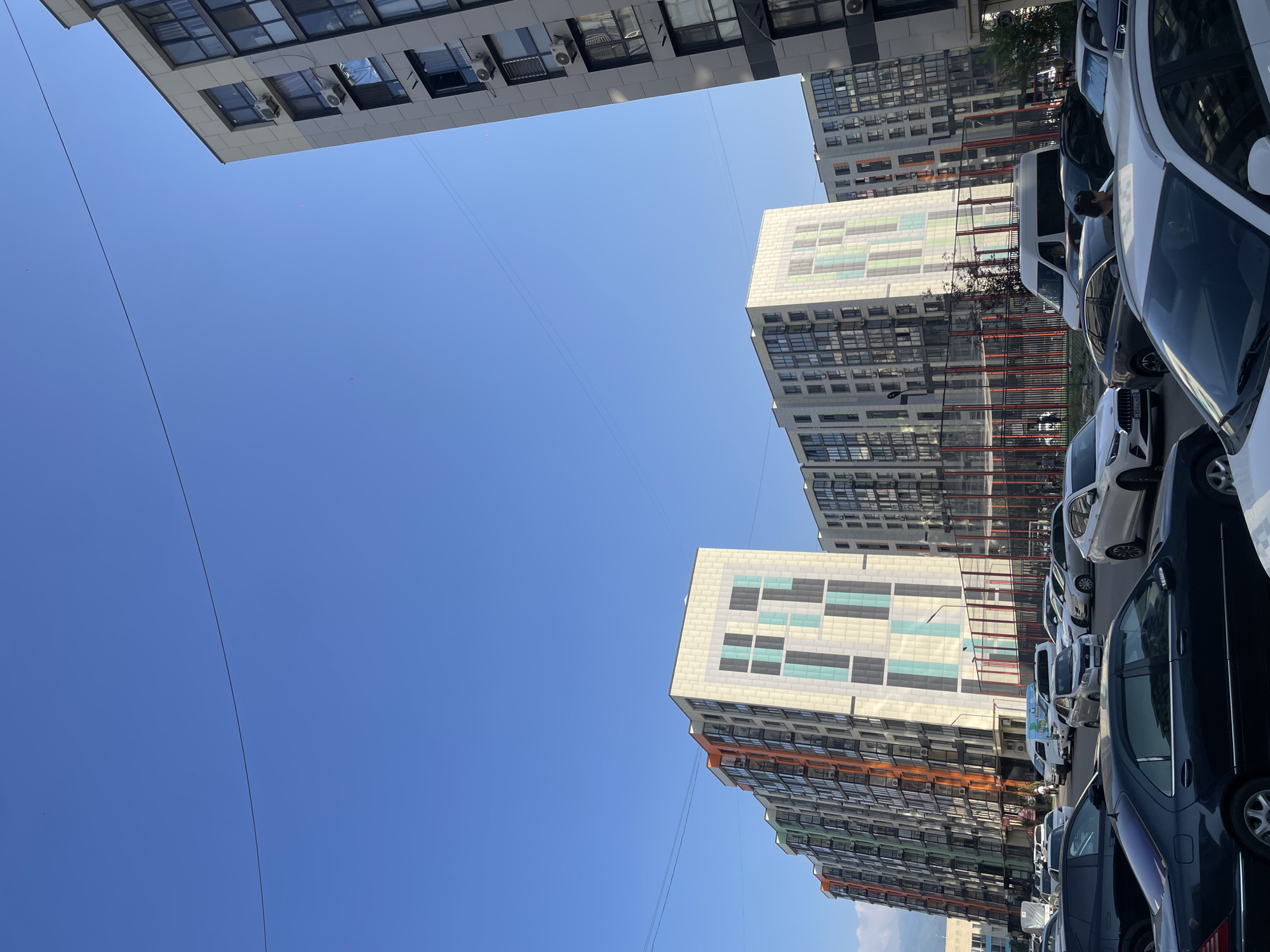 This is a photograph taken in Almaty, Kazakhstan, showing a street scene with modern apartment buildings and parked cars. The sky is clear and blue. In the foreground, several cars are parked. The buildings are multi-story residential complexes with balconies and large windows. The architecture features colorful geometric patterns on some of the facades. A fence surrounds an outdoor recreational area, possibly a sports court, behind some of the buildings. The overall impression is a contemporary urban environment.