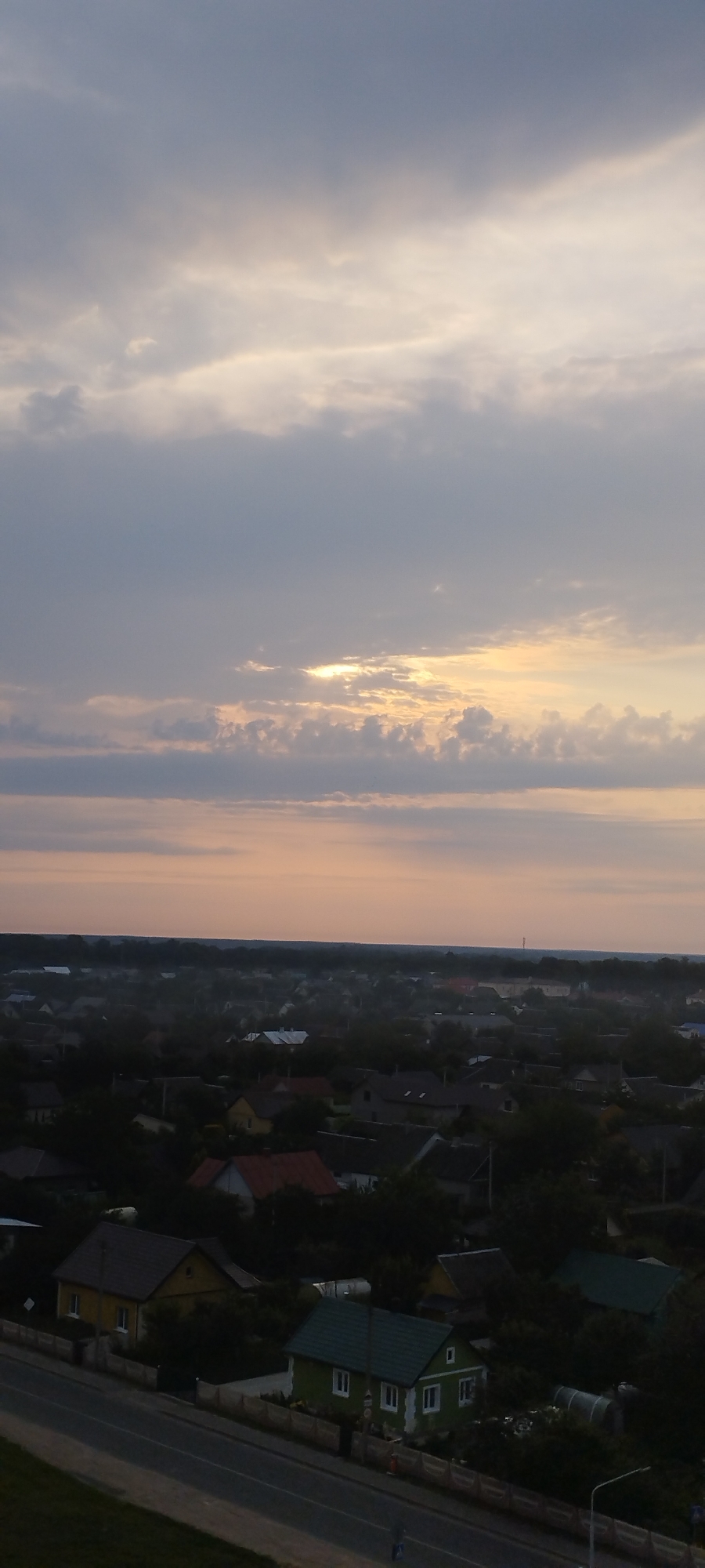 An aerial, wide shot depicts a residential area under a cloudy sky at dusk. The sky is filled with various shades of gray and pale orange clouds, with a sliver of bright sunlight peeking through in the upper half of the frame. Below the sky, a dense collection of houses with dark roofs and some scattered trees are visible. In the foreground, a paved road runs diagonally across the bottom of the image, with what appears to be a sidewalk or median strip bordering it. A few of the houses in the foreground are more clearly defined, including a yellow house with a dark roof and a green house with a dark green roof. The overall impression is that of a quiet, suburban landscape at the end of the day.