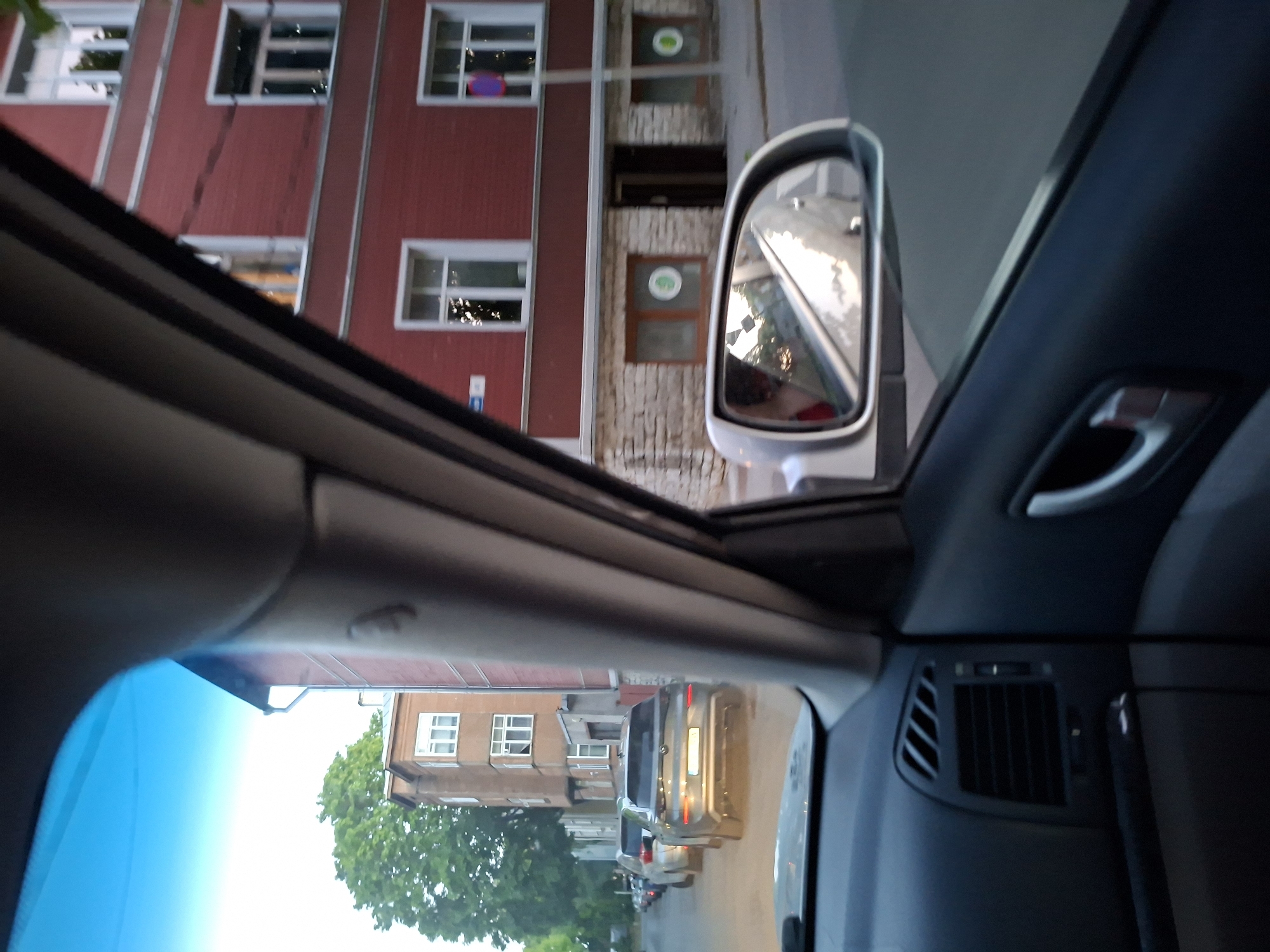 The image captures an urban street scene from the interior of a vehicle, looking towards the passenger side. The time appears to be late afternoon or early evening, characterized by bright but low-angle sunlight.
In the foreground, the dark dashboard of the car is visible, along with air vents on the right and part of a door handle on the right side panel. A blue-tinted band, likely the top edge of the windshield or a sun visor, spans the top left. The side mirror on the right reflects a portion of the vehicle's exterior and the street scene opposite, including a faint reflection of a person (presumably the driver) wearing a red garment.
The street is a narrow, unpaved or lightly paved surface with several cars parked bumper-to-bumper on the left. The closest identifiable parked car is a dark grey SUV, possibly a Volkswagen ID.4 or similar electric model, seen from the rear. Beyond the parked cars, a light brown multi-story building with white-framed windows is visible, partially obscured by trees.
Dominating the right side of the exterior view is a three-story building with a distinctive red facade and white-framed windows. Its ground floor features a stone-like base, two windows with identical green circular signs (white circles with a green border and an abstract white symbol inside), and an open doorway to the right. A blue street sign, largely illegible, is affixed to the red building's facade, to the left of a tall pole bearing a circular purple and red sign (possibly a traffic restriction sign). The architecture, including the red-facade building with its stone base and the older, light-brown building, is characteristic of renovated residential areas in Tallinn, Estonia, such as those found in Kalamaja, Pelgulinn, or certain parts of the Kesklinn district, suggesting a quieter, residential street.