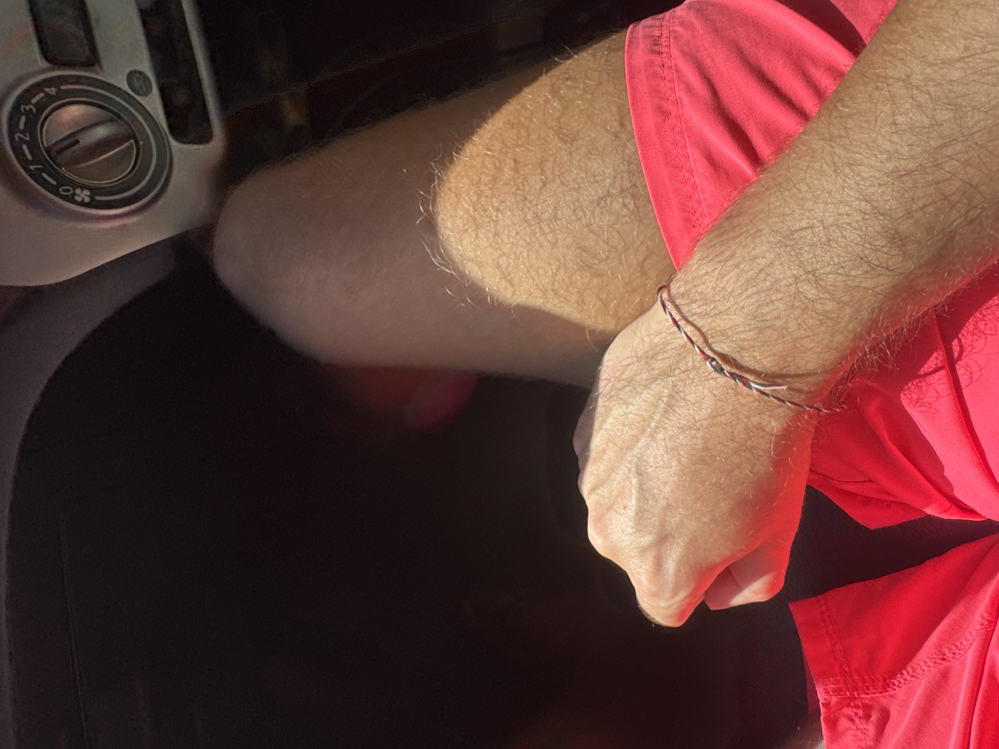 A close-up, high-angle shot shows a portion of a vehicle's dashboard, a person's hairy arm, and a red garment. The dashboard, located on the left side, features a circular vent control with numbered settings. The arm, displaying fair skin and blond hair, rests in the foreground. The person wears a multi-colored string bracelet. The red garment rests against the arm. The background appears dark. The exact location within Sukawati, Indonesia, cannot be determined from this image.