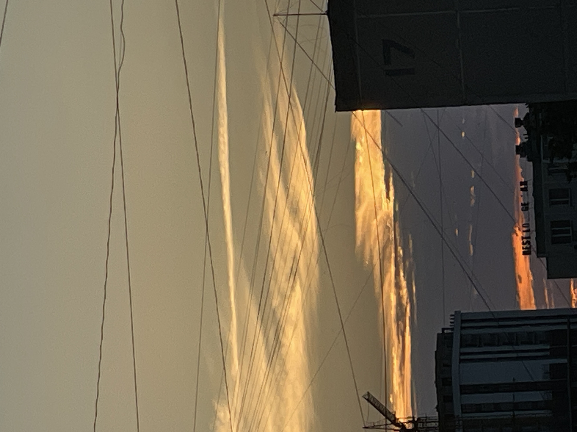 This is a vertical, low-angle shot of a cityscape at sunset. The sky is a gradient of pale yellow and orange, with streaks of light creating a dramatic effect. Multiple thin, dark wires crisscross the frame, some appearing taut and others slack. In the background, a large, dark, rectangular structure is visible, with the number "17" painted on its side. To the right, the sun reflects off glass windows of buildings, creating bright vertical streaks of orange and gold light. Another building, with horizontal bands of windows and dark framing, is situated in the lower right portion of the image. A small section of a construction crane is visible in the bottom center. The lighting suggests it is either sunrise or sunset, with the warm hues indicating the former. The presence of construction elements suggests an ongoing development in the urban environment. No people are visible. The text visible is "17" and "BEST LO". The location is Aktau, Kazakhstan.