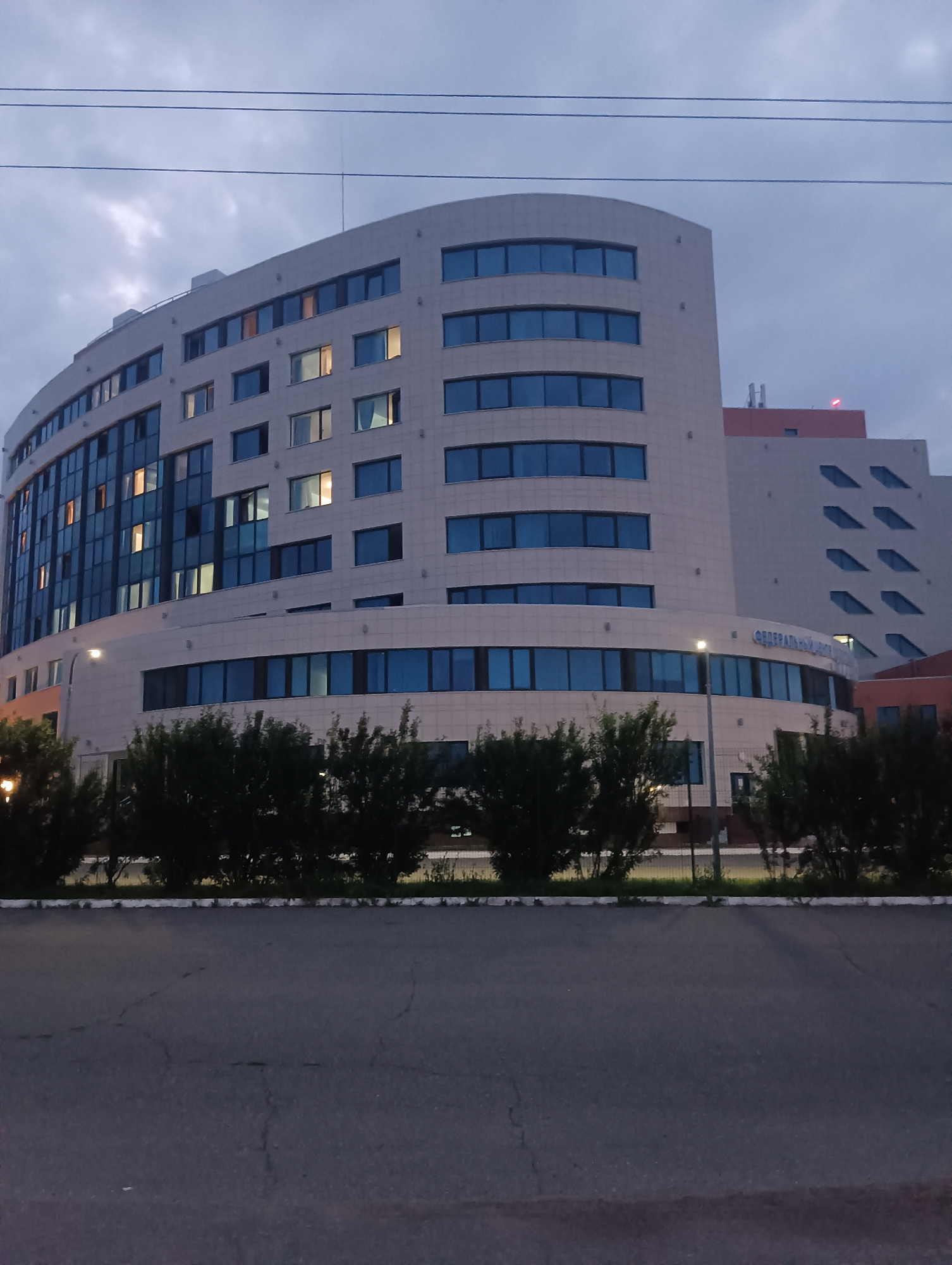 The image shows a modern, multi-story building with a curved facade, characteristic of contemporary architecture. The building is clad in light-colored panels and features a large number of windows, some of which are illuminated from within. To the right of the main building, a section of a taller structure is visible, with a distinct pattern of angled windows. The building appears to be an office or administrative complex, potentially a government or corporate facility, given the visible signage that reads "ФЕДЕРАЛЬНЫЙ" which translates to "FEDERAL". The surroundings include a paved road in the foreground and a line of dense green bushes or trees along the edge of the property. Overhead power lines are also visible against the twilight sky, which is overcast with clouds. The overall scene suggests an urban or suburban setting during the evening. The specific location in Прикамский, Russia, is identified through the visible signage and architectural style.