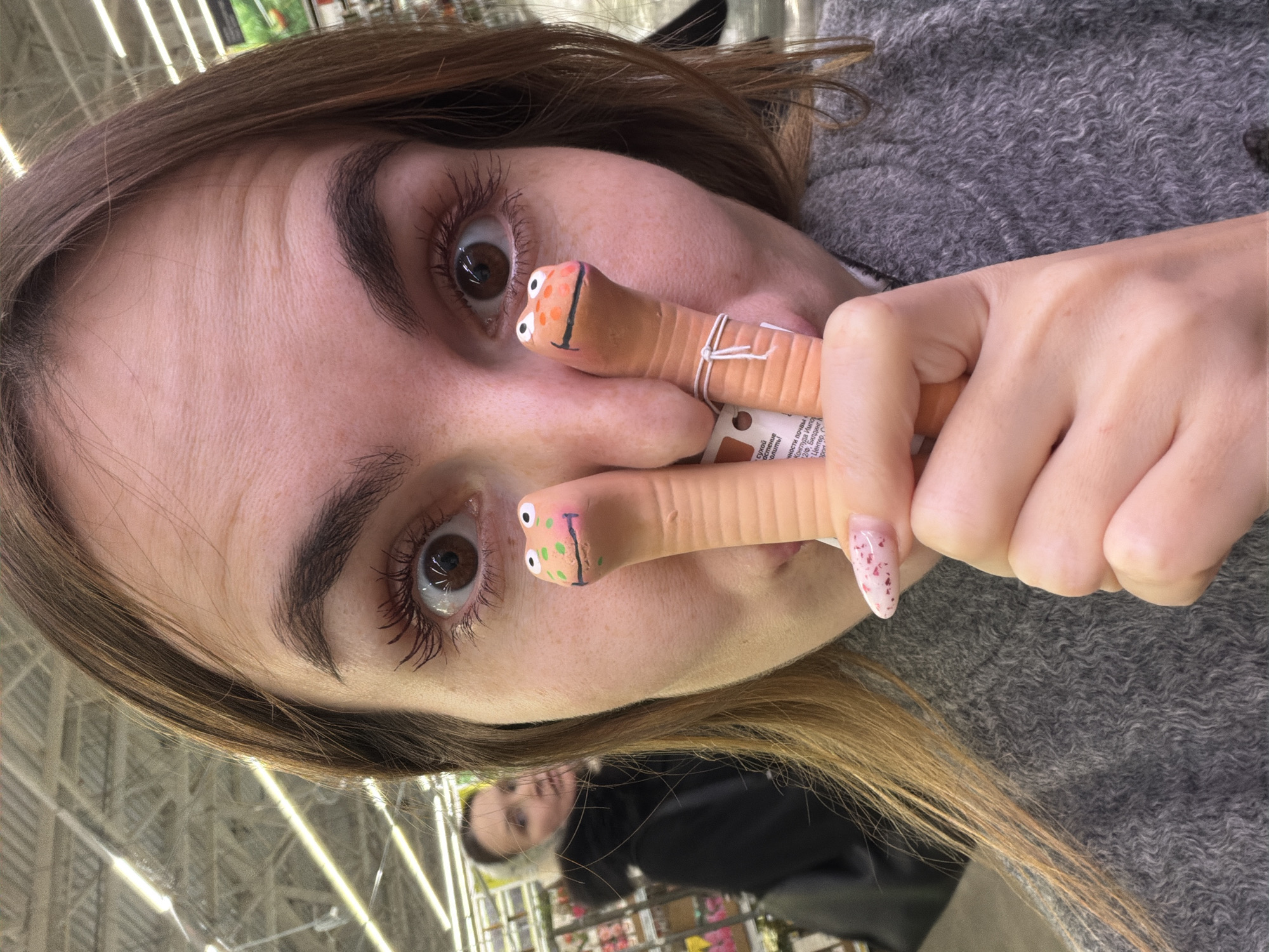 A woman's face is framed by light brown hair and she holds two toy worms up to her face, replacing her nose. Each worm has painted eyes and a painted smile. A price tag is visible on one of the worms. The woman is wearing a gray sweater. In the background, a woman is visible in a shopping cart. The location appears to be inside a store with bright overhead lighting. Text in Russian is on the price tag. сухой, растение, полить! and Центер appear. The text indicates possibly Izhevsk, Russia.