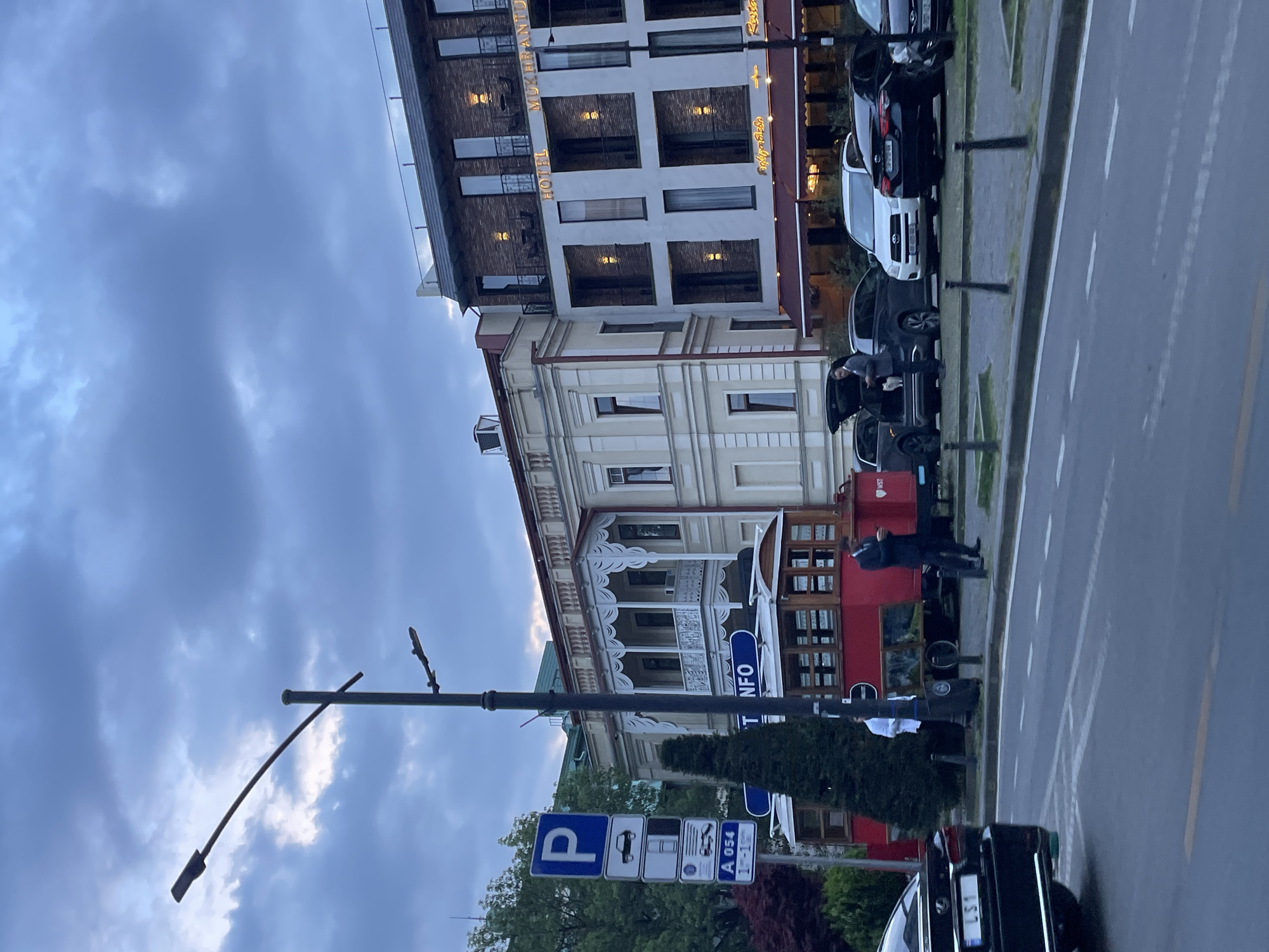 The image shows a street scene in Tbilisi, Georgia, with a view of the Hotel Mukhrantubani and another older building. Several cars are parked along the side of the road, and a person stands near the open trunk of a dark vehicle. A red structure, possibly a tram or bus stop, is visible behind the parked cars. A lamppost and parking signs are in the foreground, partially obscuring the view. The sky is overcast.
