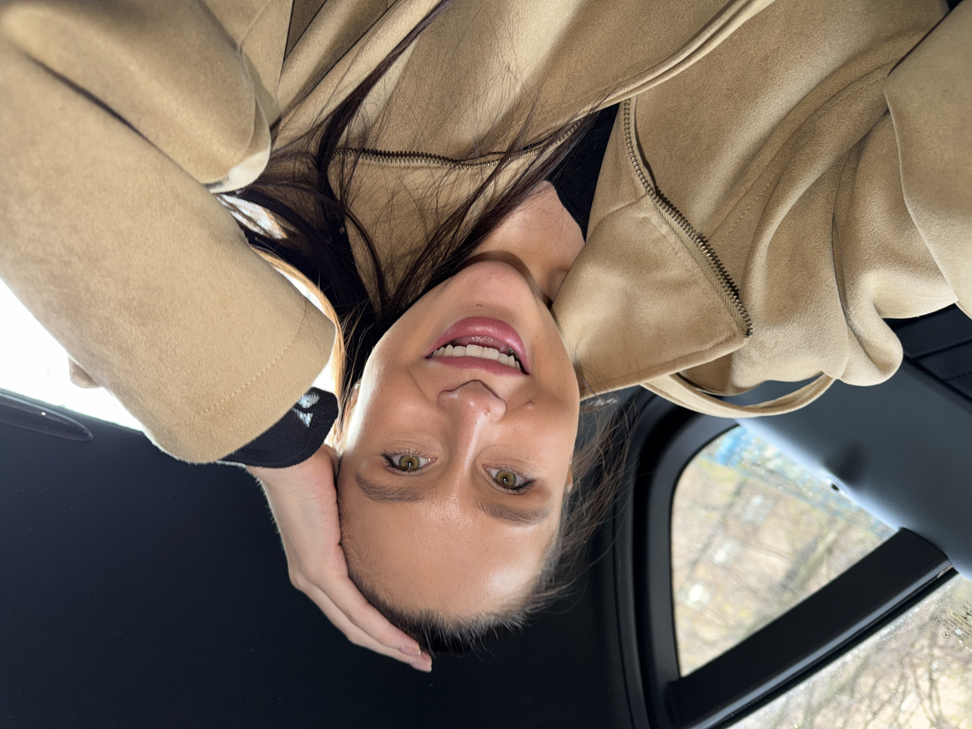 A young woman is featured in an inverted, close-up portrait, looking directly at the camera with a broad, genuine smile. Her long, dark brown hair falls around her shoulders and chest. She has bright green eyes and is wearing pink lipstick, which highlights her smile and visible white teeth. Her right hand is positioned casually against her temple and cheek.
She is dressed in a light beige or tan coat, possibly made of a soft, suede-like material, with a prominent metallic zipper running down the front. A dark, possibly black, garment is visible underneath her coat.
The setting appears to be the interior of a vehicle, suggested by the dark headliner and structural elements visible above and around her. Through a window on the right side of the frame, a blurred exterior scene is visible, dominated by bare tree branches, indicative of autumn or winter. The window also shows hints of moisture, suggesting overcast or damp weather conditions. The lighting is soft and diffused, characteristic of an indoor setting or an overcast day. No discernible text is visible within the image. This image was taken in Khimki, Russia.
