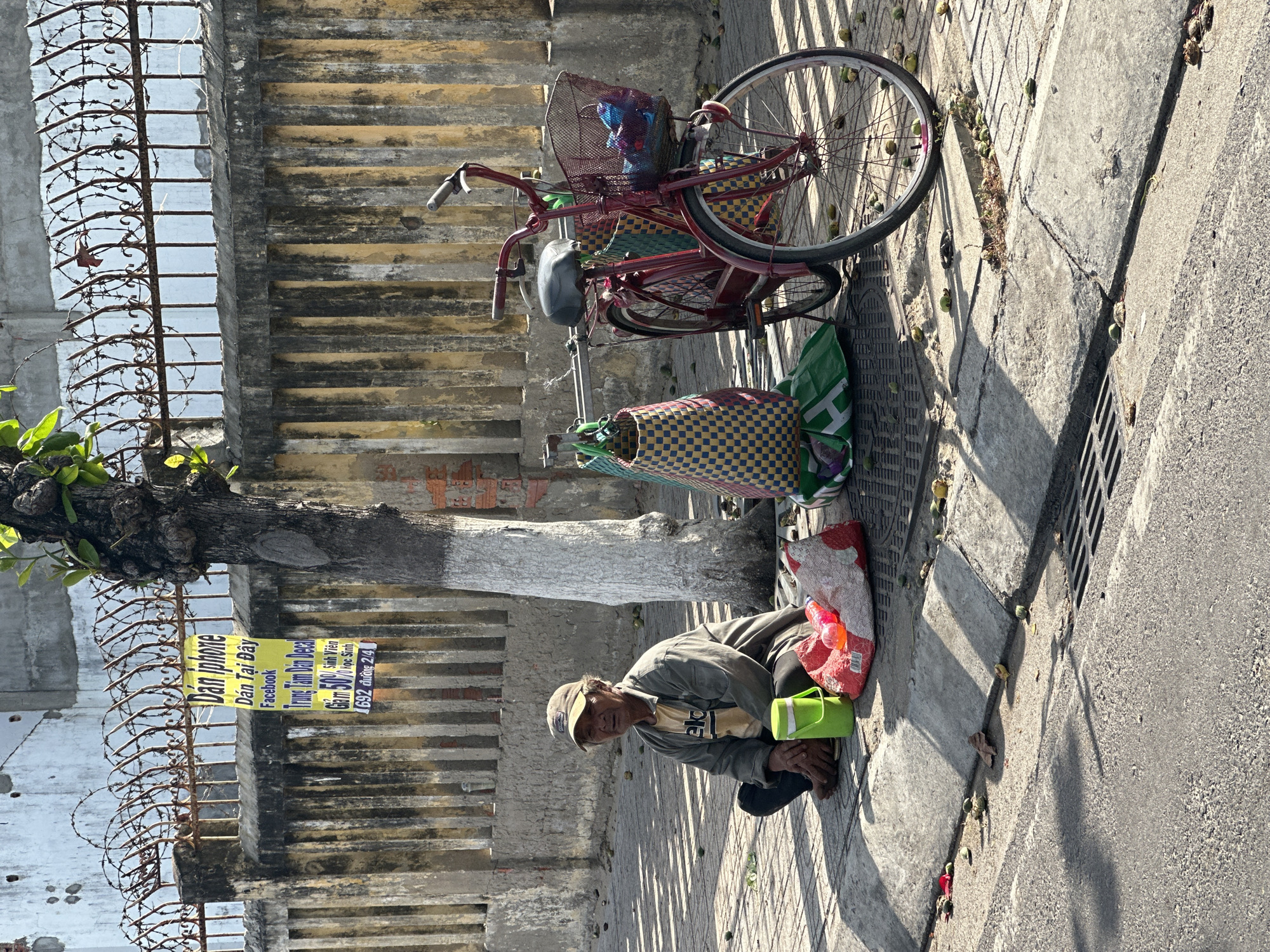 A person sits on the sidewalk in front of a fence. The person is wearing a cap and jacket and is looking at the camera. Next to the person is a green bucket, a red and white bag with a red bottle, and a bicycle with baskets. The fence is topped with barbed wire and has a yellow and black striped pattern. A sign in Vietnamese is hanging from the fence. The ground is paved with concrete slabs and there are fallen leaves and small green objects scattered around. A tree with white painted bark is next to the person. The scene appears to be a street in Bac Nha Trang Ward, Vietnam.