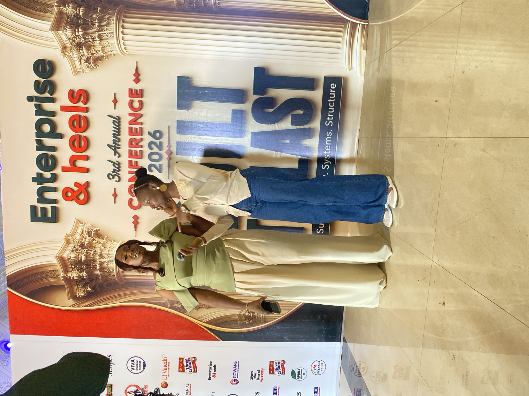 Two women are standing in front of a banner that reads "Enterprise & Heels 3rd Annual Conference 2026". The woman on the left is wearing a green off-the-shoulder top and beige wide-leg pants. She is holding a smartphone in her right hand and gesturing with her left. The woman on the right is wearing a cream-colored cardigan and dark blue jeans. She has her right hand raised to her mouth and appears to be speaking. The banner features images of ornate columns. The event appears to be taking place indoors, possibly in a conference hall. The floor is tiled. The presence of the "Enterprise & Heels" banner strongly suggests this event is related to women's entrepreneurship and professional development. While the image does not explicitly state the city of Jos, Nigeria, the logo of "Three Crowns" milk and other Nigerian brands visible on the banner suggest the event is likely taking place in Nigeria, where "Three Crowns" is a popular brand.