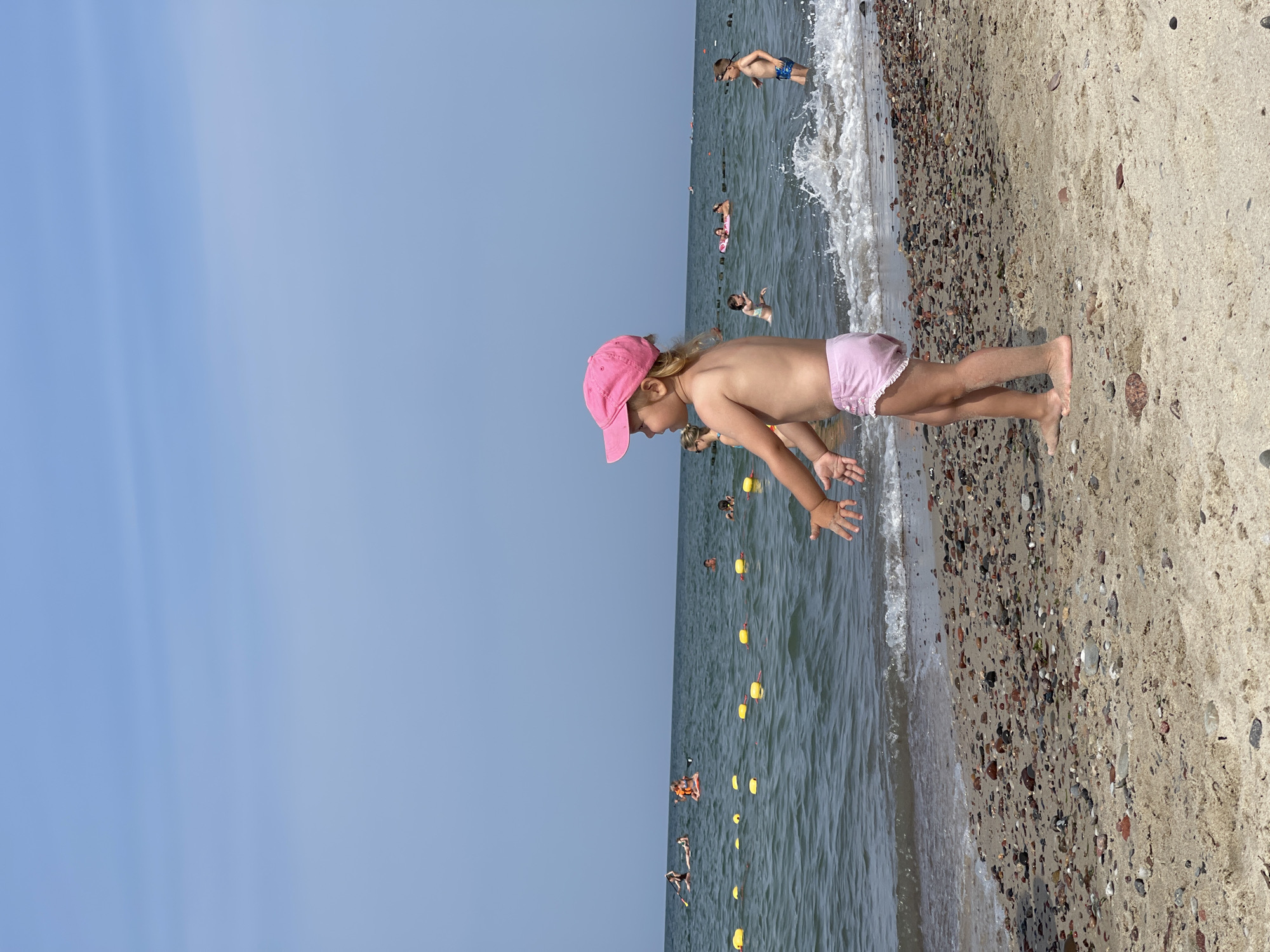This is a photograph taken on a sunny day at the beach in Zelenogradsk, Russia. In the foreground, a young girl wearing a pink baseball cap and pink bottoms stands on the sandy shore, facing the water with her arms outstretched. The beach is covered in sand and small pebbles. The sea is calm with small waves breaking near the shore. In the background, several people are visible in the water, some swimming and others on paddleboards. Yellow buoys are floating in the water, marking a boundary. The sky is clear and light blue.