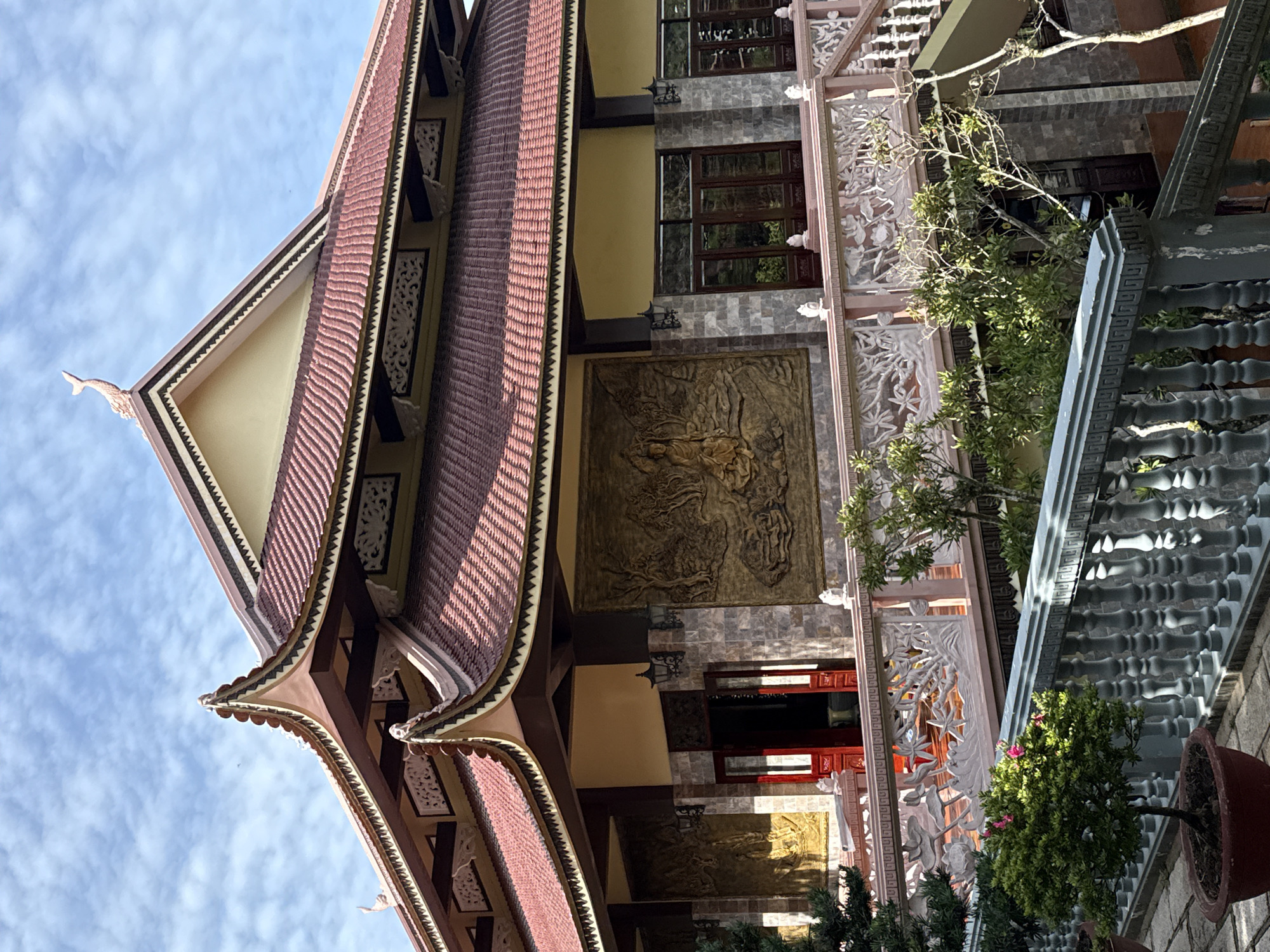 The image shows a close-up view of a traditional Vietnamese temple in Da Lat, Vietnam. The temple features a red tiled roof with ornate eaves, a yellow facade, and intricate relief carvings on the walls. A stone balustrade with decorative carvings runs along a walkway in front of the building, with lush green trees and potted plants visible. The sky above is a bright blue with scattered white clouds. The overall impression is one of serenity and architectural beauty.