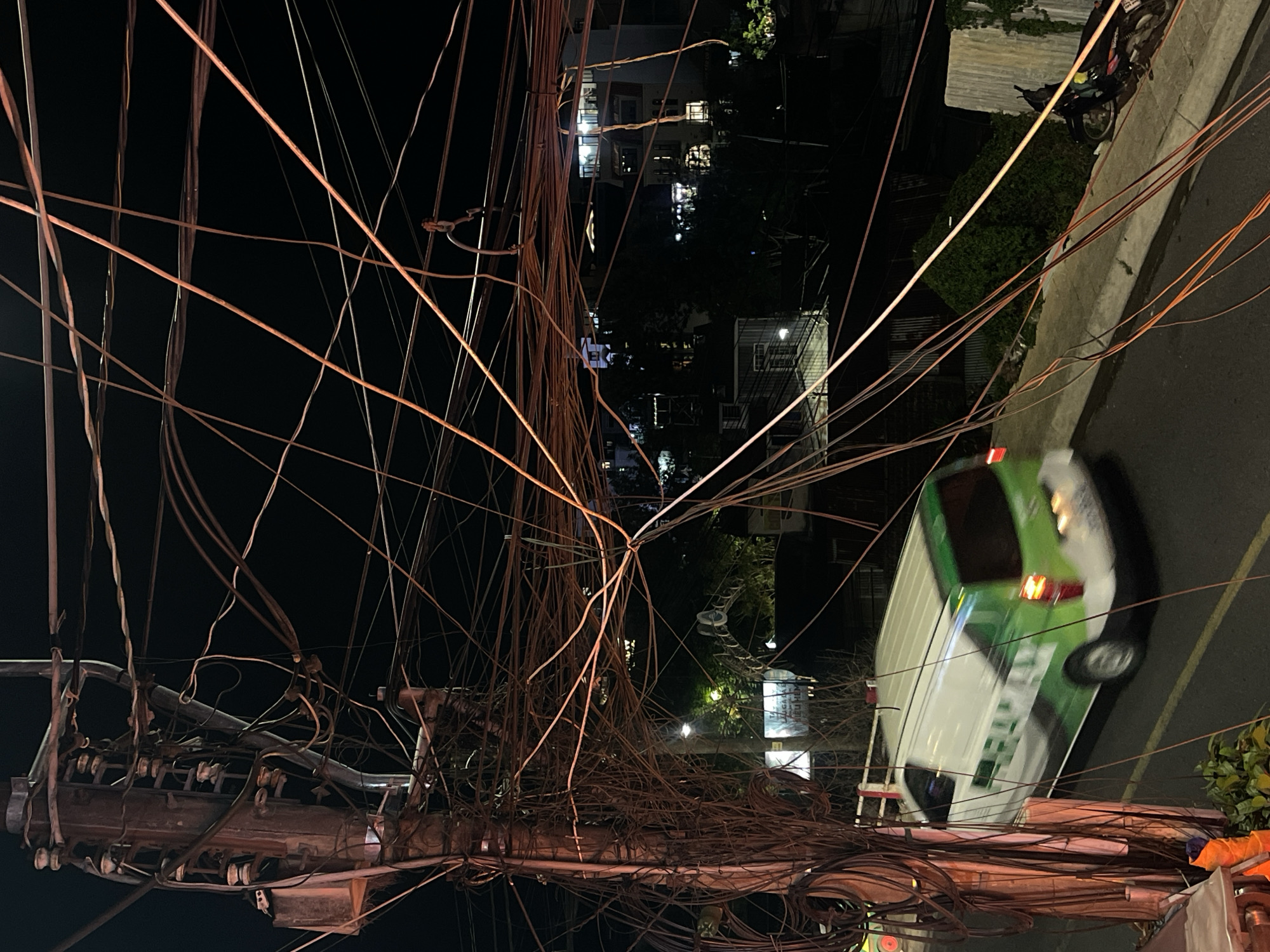 The image captures a chaotic scene of electrical wires tangled above a street in Da Lat, Vietnam at night. A utility pole laden with wires is prominently visible in the foreground. Multiple thin, dark wires crisscross the sky, obscuring the buildings and streetlights visible in the background. A white and green van is parked on the street, with its brake lights illuminated. The road is made of concrete and runs down toward the right of the image. Vegetation is visible beside the vehicle.