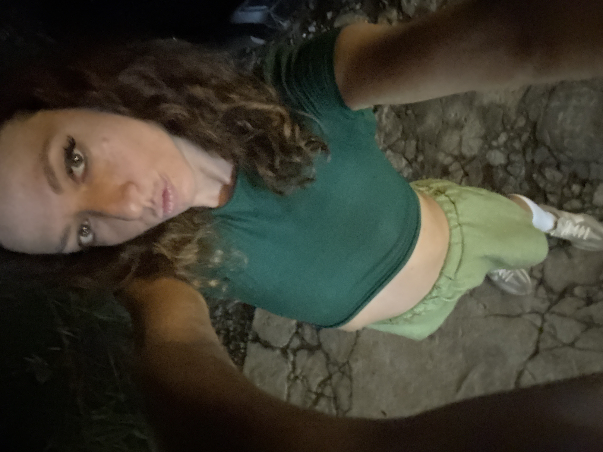 A woman with curly brown hair is taking a selfie from a high angle. She is wearing a green crop top and light green shorts. One of her arms is visible at the bottom of the frame, and another extends to hold the camera. She is standing on pavement with cracks. White socks and sneakers are visible on one of her feet. Based on the cracked pavement it's possible the location is 1 Ward, Hà Giang City, Hà Giang, Vietnam.