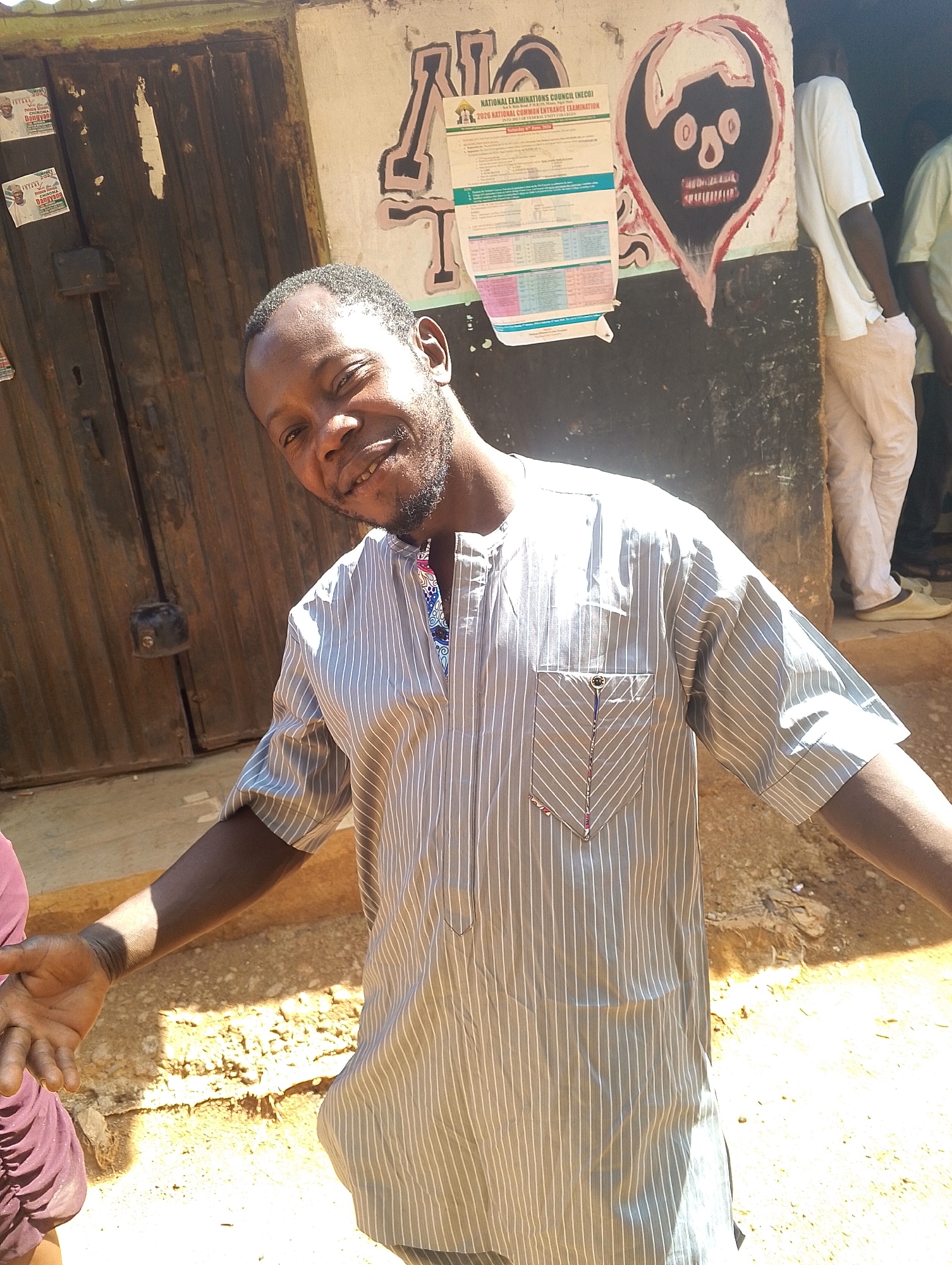 The image depicts a man, possibly in his late 20s or early 30s, standing outdoors on what appears to be a sunny day. He has dark skin, short hair, and a beard. He is wearing a light gray, vertically striped shirt with a pocket. His arms are outstretched, and he is smiling at the camera.
Behind him is a weathered wooden door and a wall with some posters and graffiti. One poster has the words "NATIONAL EXAMINATIONS COUNCIL (NECO) 2026 NATIONAL COMMON ENTRANCE EXAMINATION" visible. The graffiti includes a crude drawing of a face with large eyes and a red mouth.
To the right of the man, partially visible, are other people dressed in casual clothing. The ground appears to be sandy.
Given the presence of NECO exam posters, it can be inferred that the location may be near a school or an examination center, potentially in the city of Jos, Nigeria.