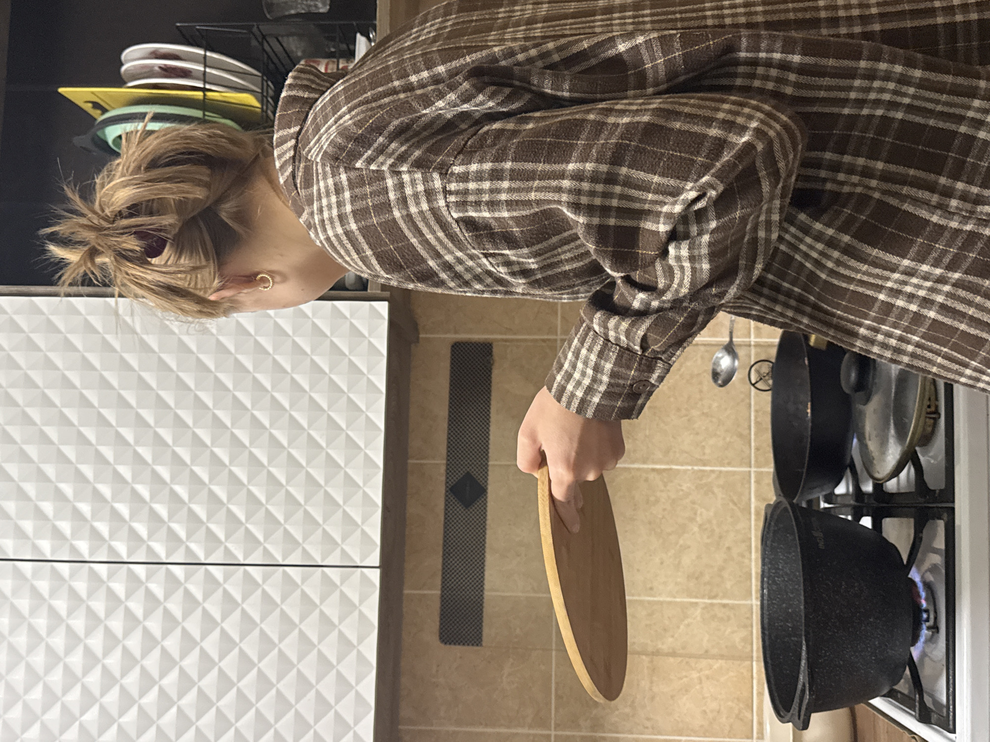 An indoor, horizontally oriented image shows a person in a kitchen, engaged in cooking activities. The person, viewed from the back and side, wears a brown and off-white plaid long-sleeved shirt. Their hair is gathered in a messy bun, and a small gold hoop earring is visible on their right ear.
The person is holding a light brown wooden circular board or lid in their right hand, positioned near a gas stove. On the stove, two dark-colored pots or cauldrons are heating, with a visible blue flame under the pot closest to the viewer. A metal spoon is resting near the other pot.
The kitchen features white cabinets or drawers with a textured, geometric pattern on the left side of the frame. Above these cabinets, a black wire rack holds several stacked plates and bowls in various colors (white, yellow, green). The floor or wall area between the cabinets and the stove is covered in light brown square tiles. A dark, patterned rectangular mat is visible on the tiled surface.
The scene depicts a domestic setting, likely in Chaikovsky, Russia, focused on meal preparation. No specific time of day or emotional state is discernible. There is no clearly legible text visible in the image.