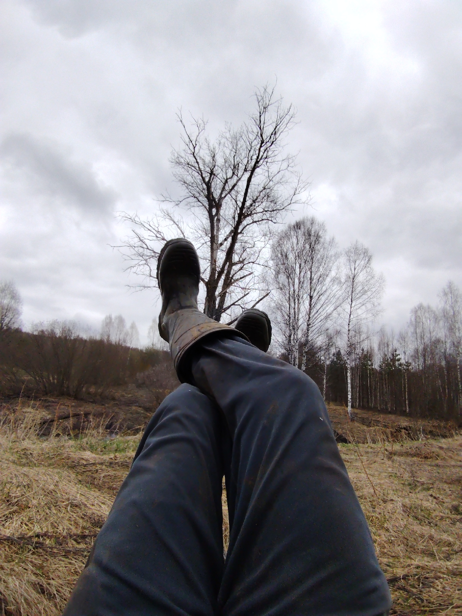 The image shows a first-person perspective of someone lying down outside in Markovskii, Russia. The person is wearing dark pants and rubber boots with leather straps. The feet are crossed. The ground is covered in dry grass. In the background is a sparse forest with leafless trees under an overcast sky.