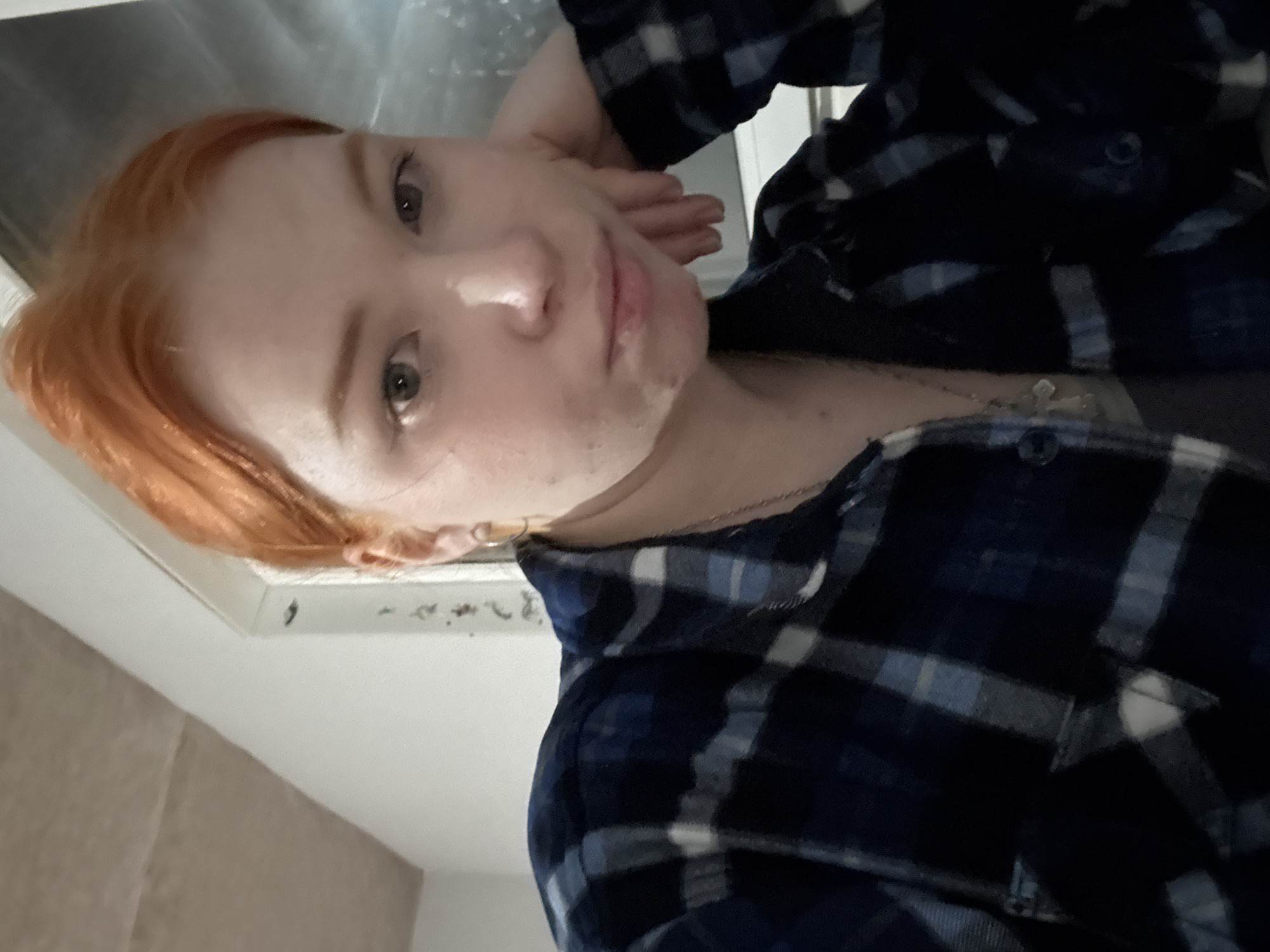 A close-up portrait shows a person with reddish-orange hair and fair skin, looking directly at the camera. The image appears to be taken indoors, oriented vertically but currently rotated counter-clockwise. The person is depicted from the chest up, wearing a dark blue and light-colored plaid button-up shirt. A thin silver chain necklace with a small, light-colored pendant, possibly a cross, hangs around their neck. A small hoop earring is visible in their right ear. One hand is gently raised and positioned near the side of their face. The background is indistinct, appearing as a light-colored wall or ceiling with some darker, textured areas, possibly a vent or fixture. The lighting creates a highlight on the person's nose and forehead. Their expression is neutral, bordering on serious or pensive. There is no visible text in the image.
