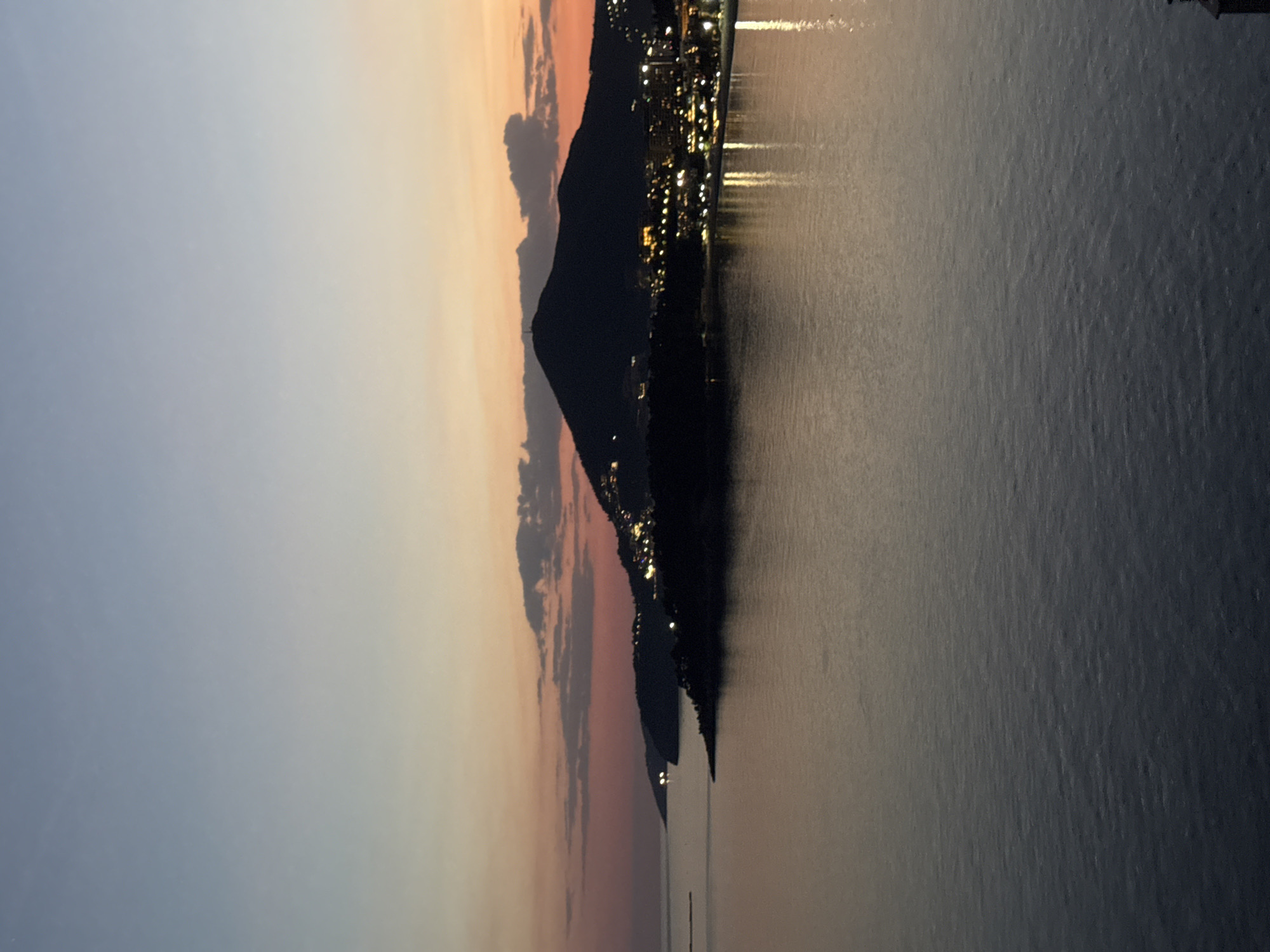 The image shows a coastal scene at sunset, likely in Rafailovići, Montenegro. A dark mountain range stands between the water and the sky. The mountain is dotted with lights, indicating a city or town. The water surface reflects the city lights. The sky transitions from light blue to orange and pink at the horizon, indicating sunset. Clouds are visible near the mountain range.