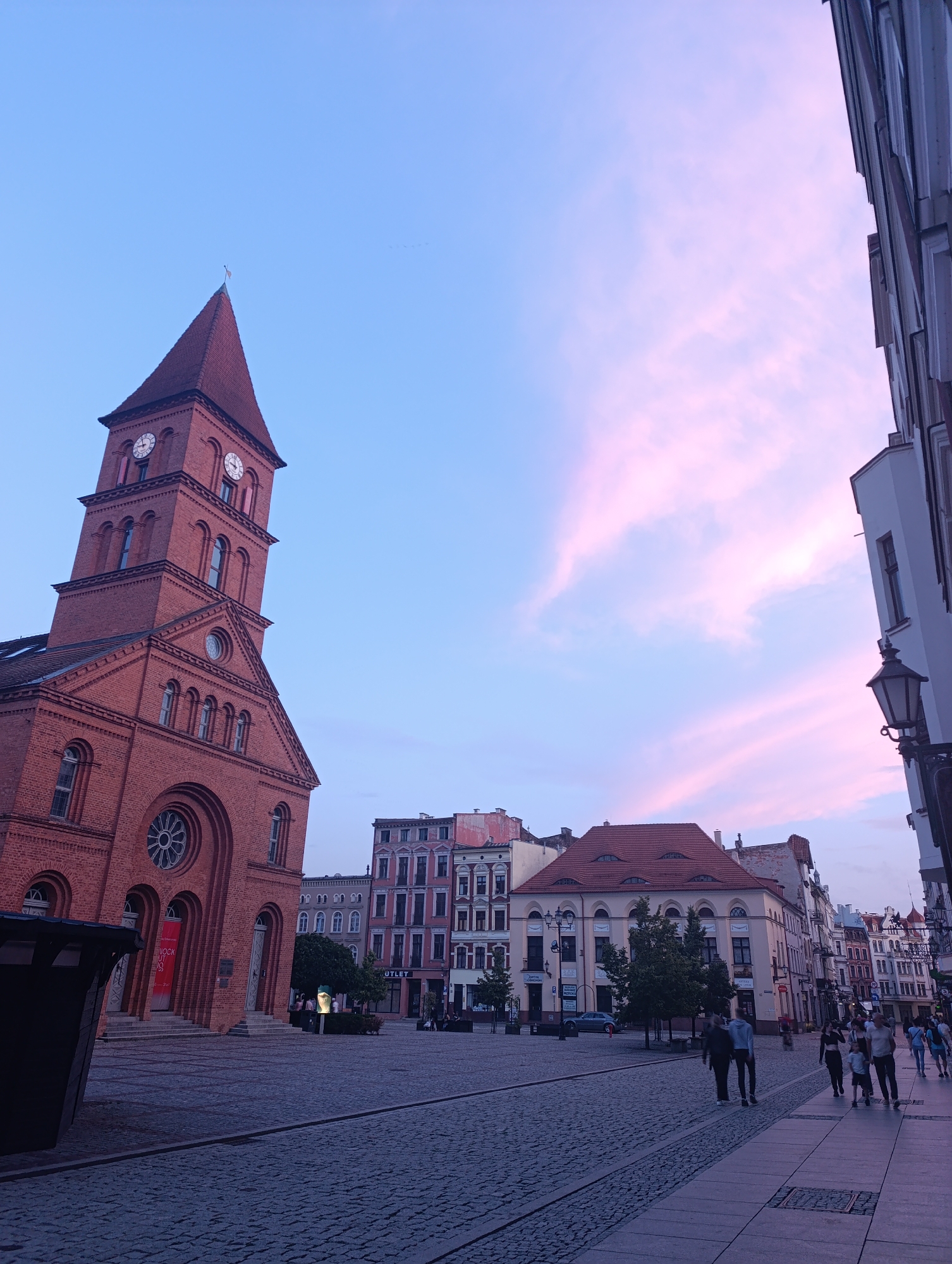This image depicts a view of the Old Town Square in Toruń, Poland, at dusk. The sky is a gradient of blue with streaks of pink and purple from the setting sun, illuminating the clouds. In the foreground, a cobblestone street leads into the square. To the left, a large, red brick church with a tall, pointed steeple and visible clock faces dominates the scene. The church features arched windows and a large circular window above the entrance. Several multi-story buildings with distinct architectural styles line the square, some painted in pastel colors. On the right side, the edge of a white building with a black street lamp attached to it is visible. A group of people are walking on the sidewalk in the distance, some in pairs and others in small groups. The overall atmosphere is calm and serene, characteristic of an early evening in a European city.